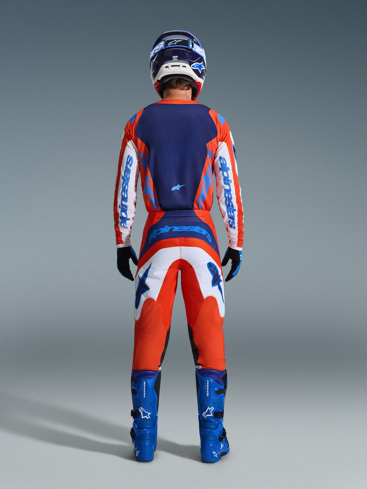 Alpinestars 2026 Fluid Grid-set, motocrossförare sedd bakifrån i en komplett racingoutfit i orange och UCLA Blue, långärmad tröja med marinblå rygg och vita ärmar, matchande orangea byxor med blå grafik, blå stövlar och matchande hjälm, professionell utrustning för terrängkörning