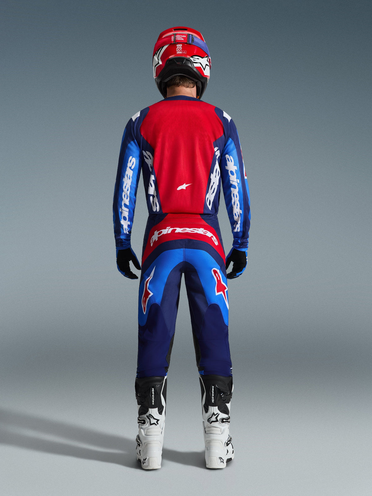Vy bakifrån på en motocrossförare iförd 2026 Fluid Grid Jersey i blått och rött, med en stor röd ryggpanel och blå ärmar med vit Alpinestars-logotyp, matchat med tillhörande byxor, vita och svarta stövlar och en röd hjälm för en komplett outfit för terrängkörning.
