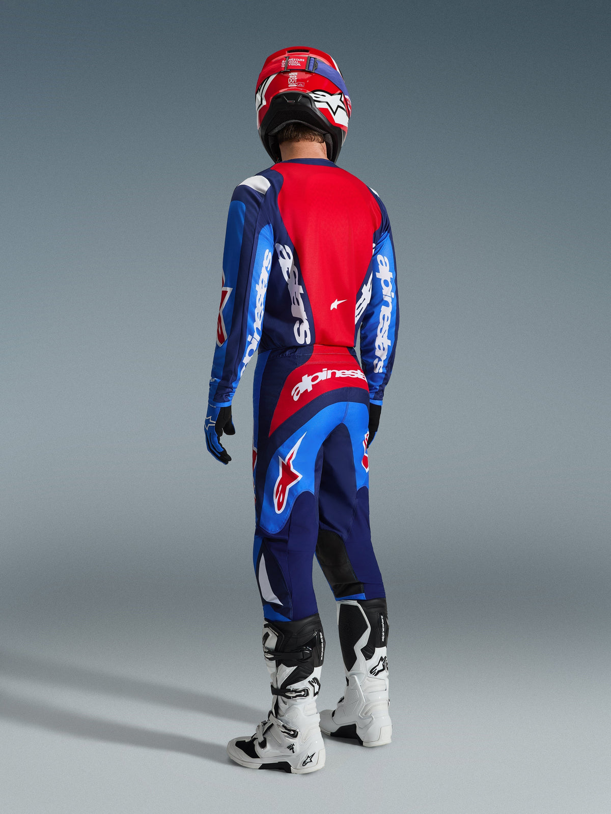 Motocrossförare sedd bakifrån klädd i Alpinestars 2026 Fluid Grid-tröja i blått och rött, matchande motocrossbyxor med tydlig vit logotyp, röd hjälm och vita och svarta stövlar, komplett utrustningsset för terrängkörning