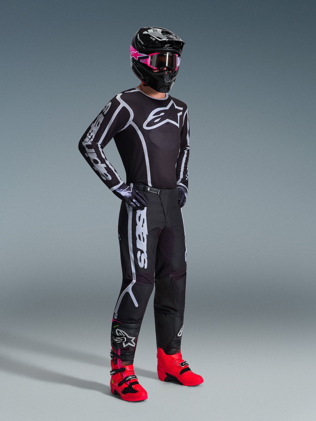 Alpinestars 2026 Fluid Apex utrustningsset, motocrossförare i svart tröja och byxor med vita och grå grafiska detaljer, svart hjälm med rosa goggles och starkt rosa racingstövlar, matchande professionell motocrossoutfit