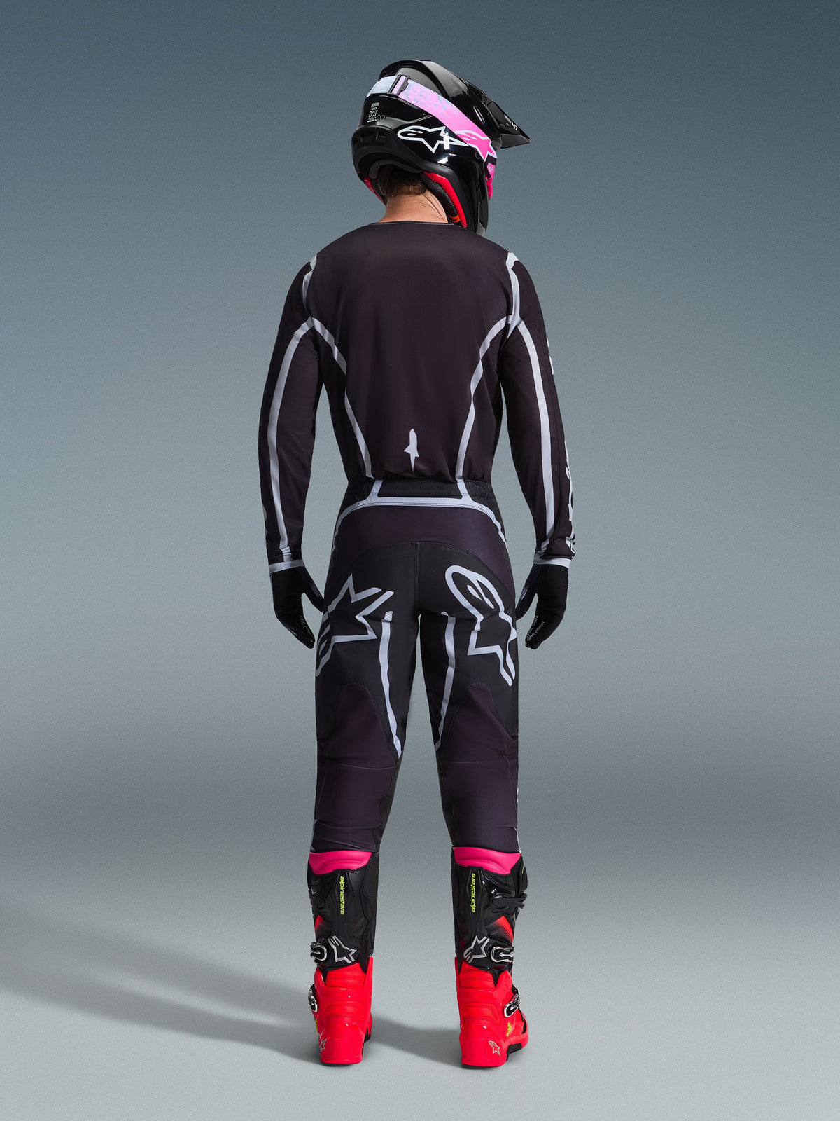 Motocrossförare sedd bakifrån bärande Alpinestars 2026 Fluid Apex Gear Set, svart tröja och byxor med grå geometriska linjer och stjärnlogotyper, svart hjälm med rosa detaljer, klarröda och svarta stövlar, matchande motocrossutrustning