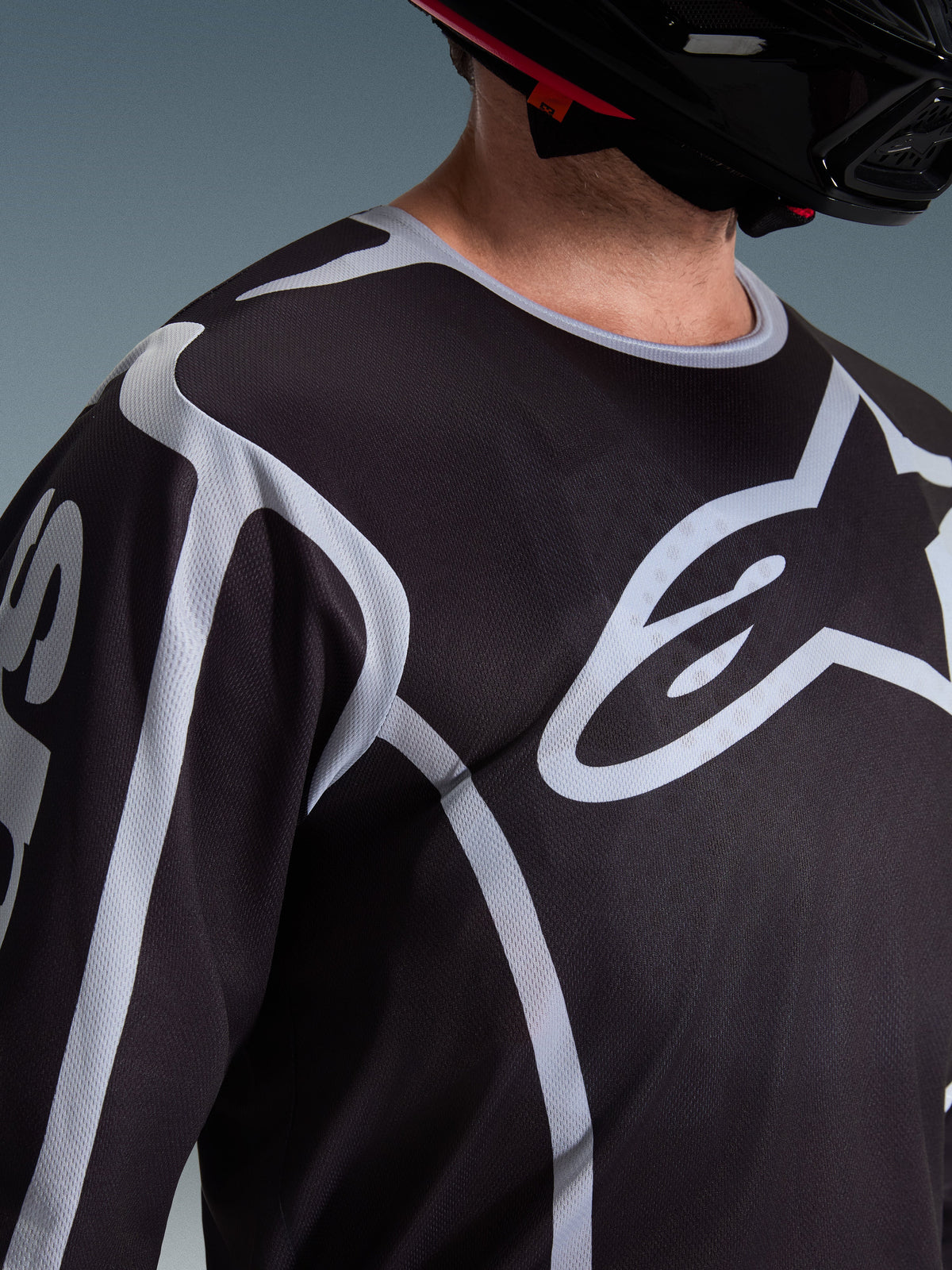 Alpinestars 2026 Fluid Apex-tröja, motocrosströja, svart och grå, med en stor Alpinestars-logotyp på bröstet, sportkrage, bärs med en svart hjälm, prestandautrustning för terrängkörning