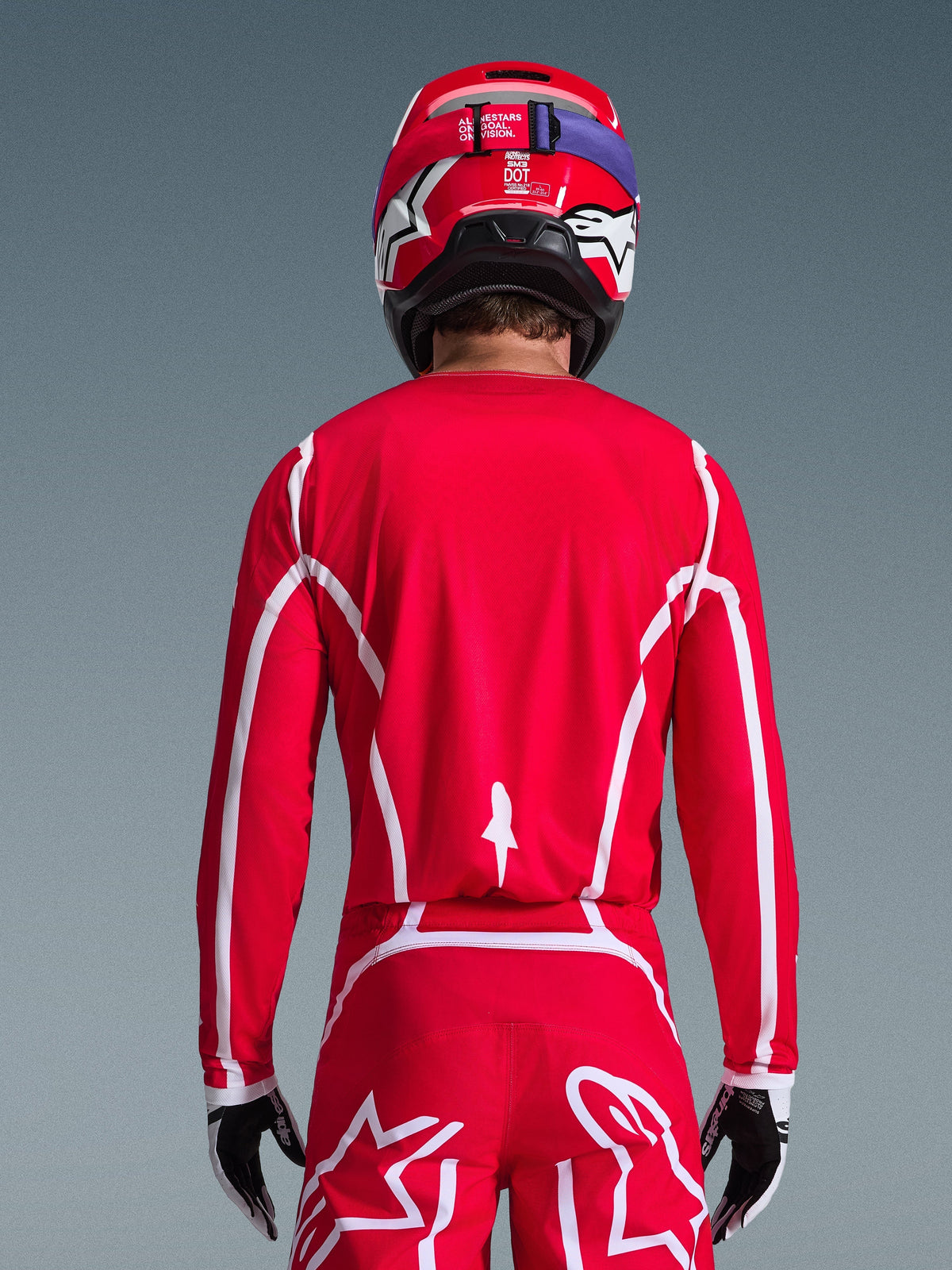 Motocrossförare sedd bakifrån i Alpinestars 2026 Fluid Apex-tröja i rött och vitt med geometriska vita mönster, stylad med matchande motocrossbyxor och en röd hjälm, komplett racingutrustning för offroad-prestanda.