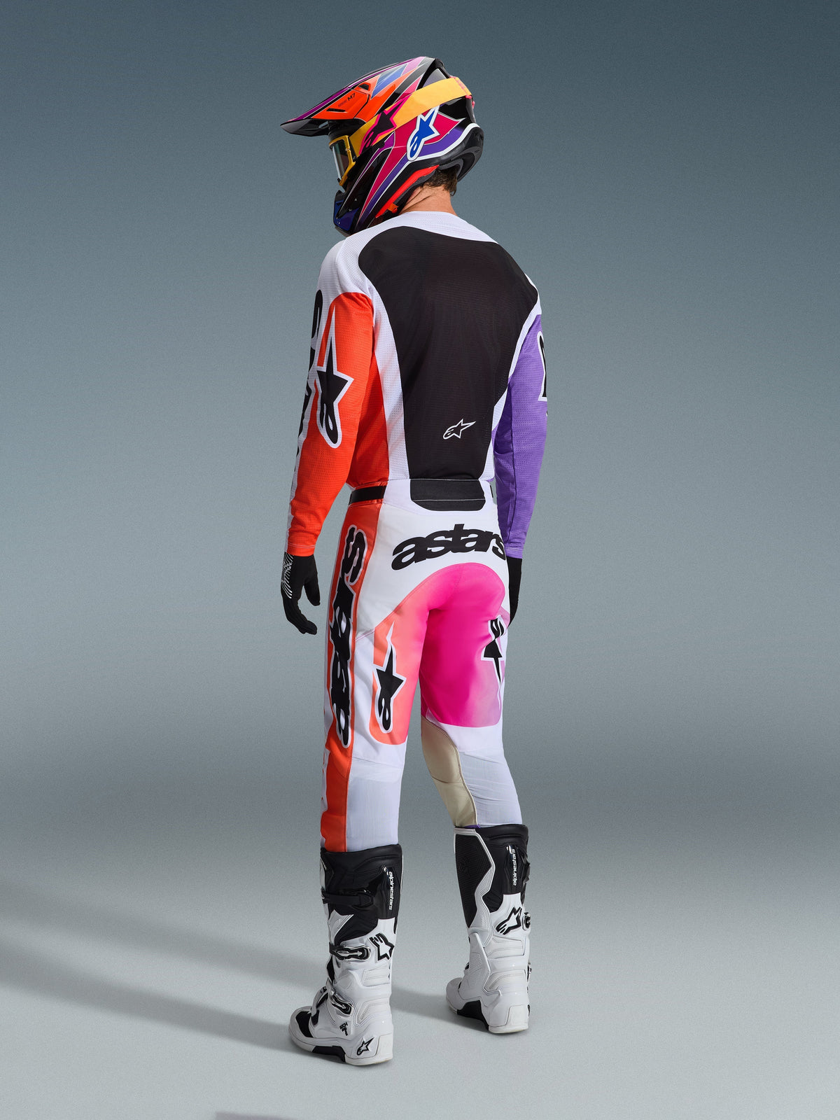 Motocrossförare sedd bakifrån bärande Alpinestars 2026 Racer Air Portl-byxor i Multicolor, med vita, orange och lila paneler, neonrosa detaljer och tydlig svart logotyp, matchande tröja, färgglad mönstrad hjälm samt vita och svarta stövlar.