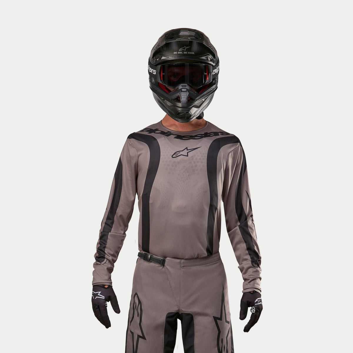 Motocrossförare iklädd Alpinestars 2024 Fluid Lurv-tröja i Mud Black, ett jordnära gråbrunt tyg med svarta grafiska ränder och en stjärnlogotyp på bröstet, kombinerat med en svart hjälm, röda goggles och svarta handskar för en koordinerad look för terrängkörning