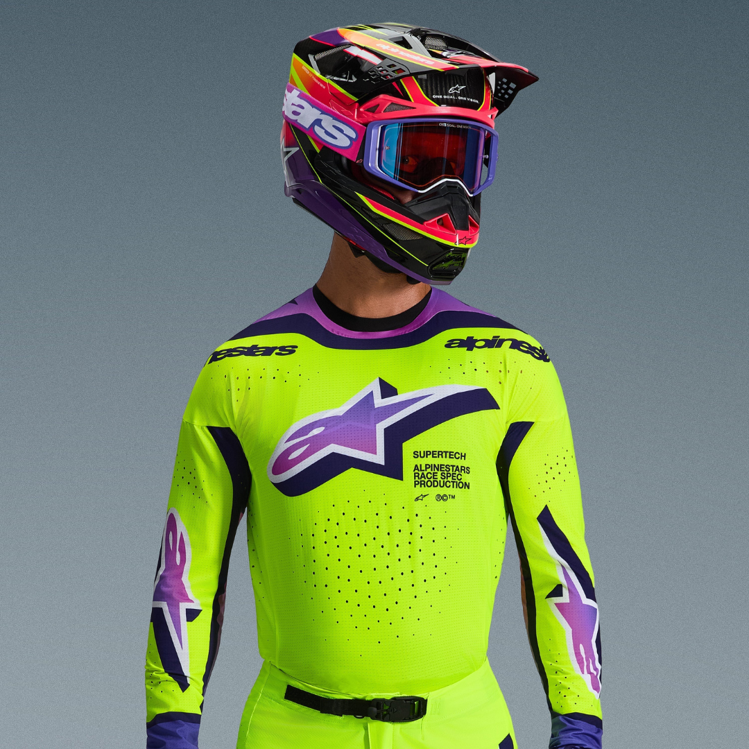 Motocrossförare i Alpinestars 2026 Supertech Vista-utrustning, neongul tröja med lila och svarta detaljer, stor lila gradientlogotyp på bröstet, matchande färgstark flerfärgad hjälm och lila goggles, visar laserskuren ventilation och professionell atletisk passform