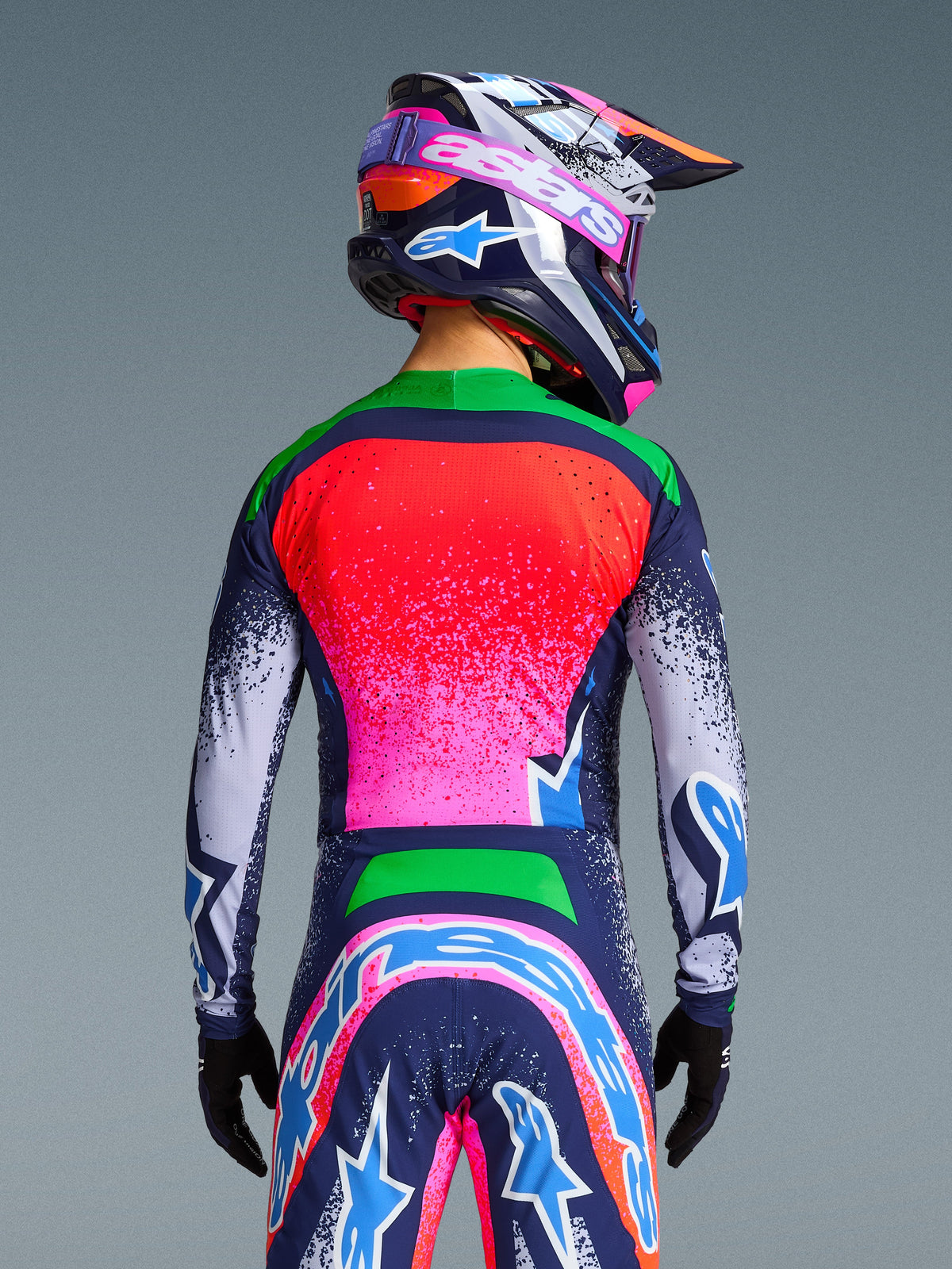 Alpinestars Limited Edition Supertech Records-tröja, ryggvy av en motocrossförare i ett färgstarkt flerfärgat ställ med rosa och röd gradient på ryggen, gröna axlar och marinblå spräckliga ärmar, kombinerat med matchande hjälm och byxor för en professionell racingstil.