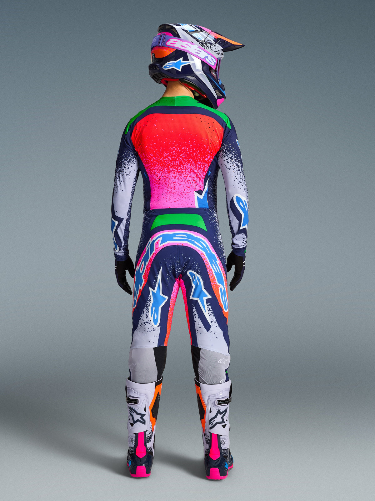 Motocrossförare sedd bakifrån i Alpinestars Limited Edition Supertech Records Gear Set, färgstark multifärgad tröja och byxor med splatterdesign i rosa, orange och blått, matchande hjälm, svarta handskar och vita och rosa stövlar, professionell motocrossutrustning.