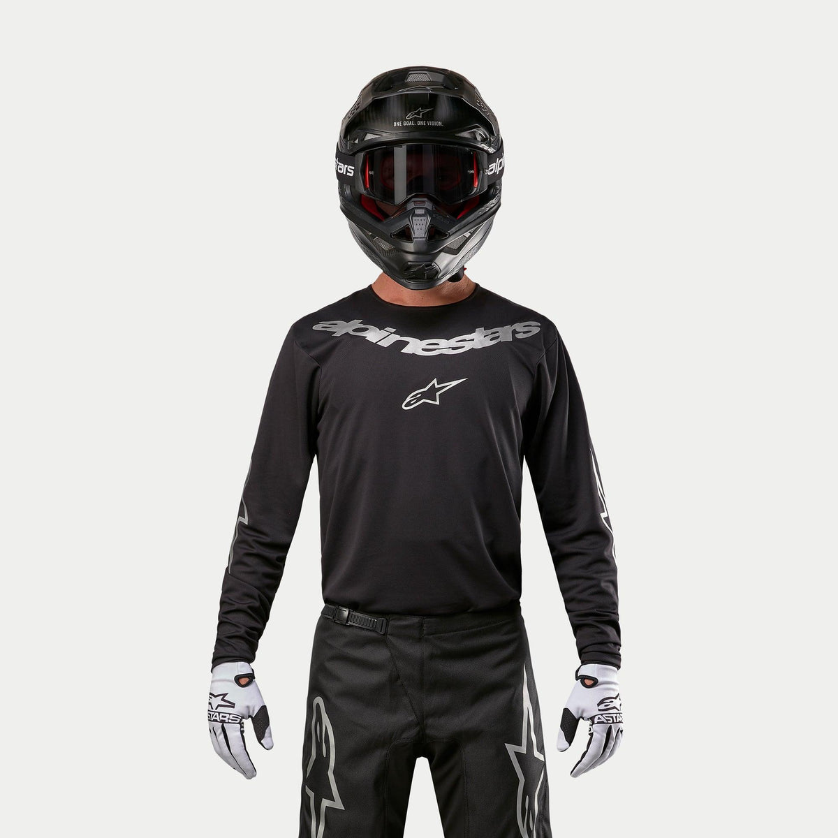 Alpinestars 2024 Fluid Graphite Jersey, svart och silverfärgad motocrosströja, bärs av en förare med svart hjälm och röda goggles, silverfärgad Alpinestars-logotyp på bröstet, matchad med svarta byxor och vita handskar för terrängkörning