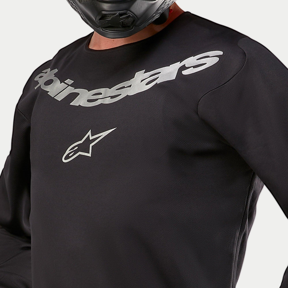 Alpinestars 2024 Fluid Graphite-tröja, motocross-tröja, svart och silver, närbild av bröstpartiet med Alpinestars-märkning och stjärnlogotyp i silver, konstruktion i andningsbart poly-material, bärs av en förare i svart hjälm