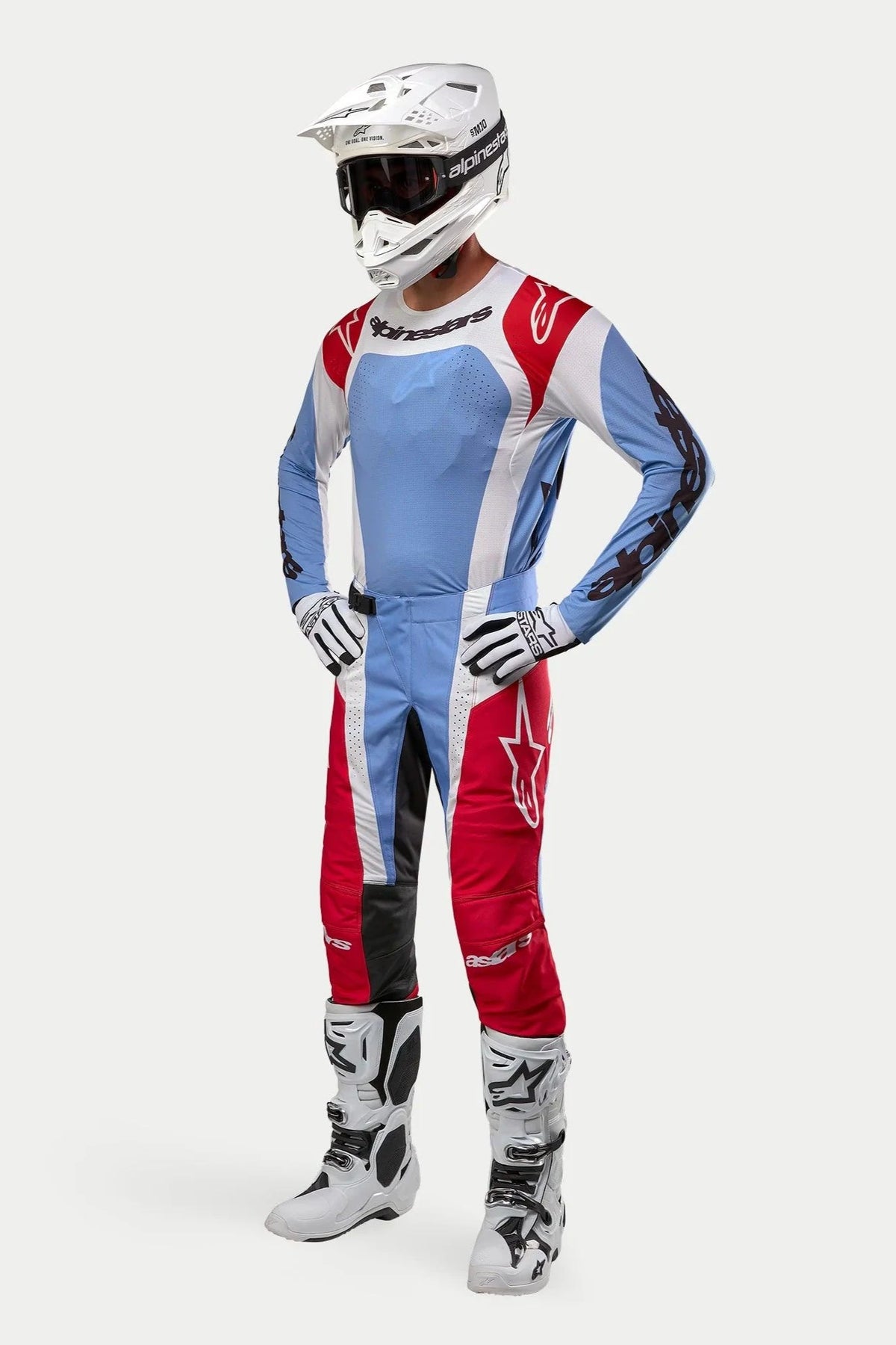 Motocrossförare i komplett racingutrustning, Alpinestars 2024 Techstar Ocuri-tröja i ljusblått, mars-rött och vitt, matchande byxor, vit hjälm med goggles, vita stövlar och vita och svarta handskar.