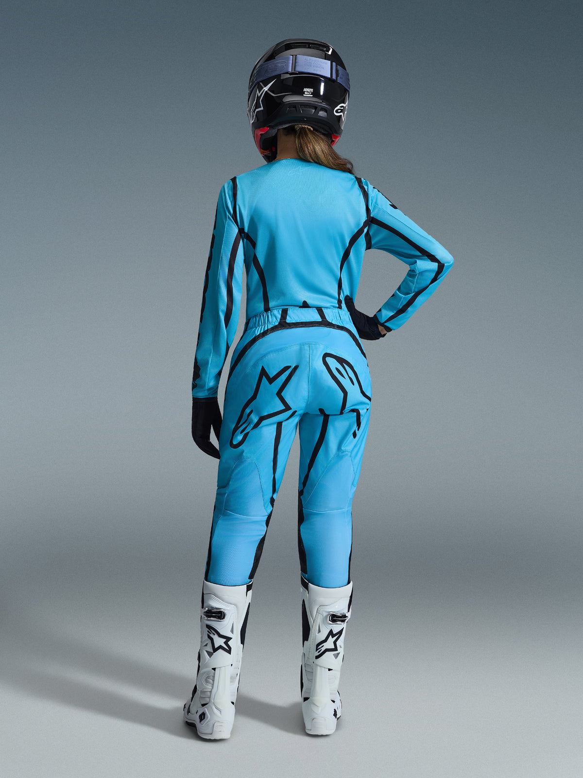 Female motocross rider from the back, wearing the Alpinestars 2026 Woman Stella Fluid Apex Gear Set in bright blue with black graphic line accents, featuring a large black star logo on the pants, paired with white and black boots and a black helmet.