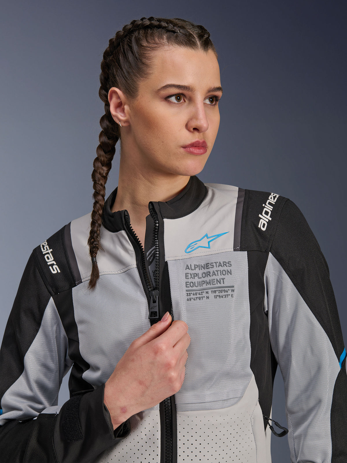 Kvinna i Alpinestars Stella ST-2 Air Jacket i Ice Gray och svart, närbildsporträtt, med meshpaneler för ventilation, 'ALPINESTARS EXPLORATION EQUIPMENT'-märkning på bröstet med en blå logotyp, modern äventyrsmotorcykeljacka