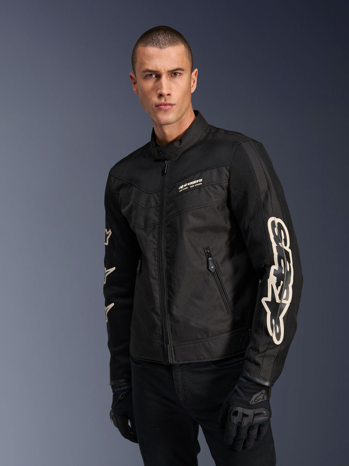 Alpinestars T-Dyno Air Jacket in Black, male model wearing a vintage-inspired textile motorcycle jacket with mesh panels for ventilation, large white Alpinestars logo on sleeves, paired with black riding gloves, urban motorcycle gear design