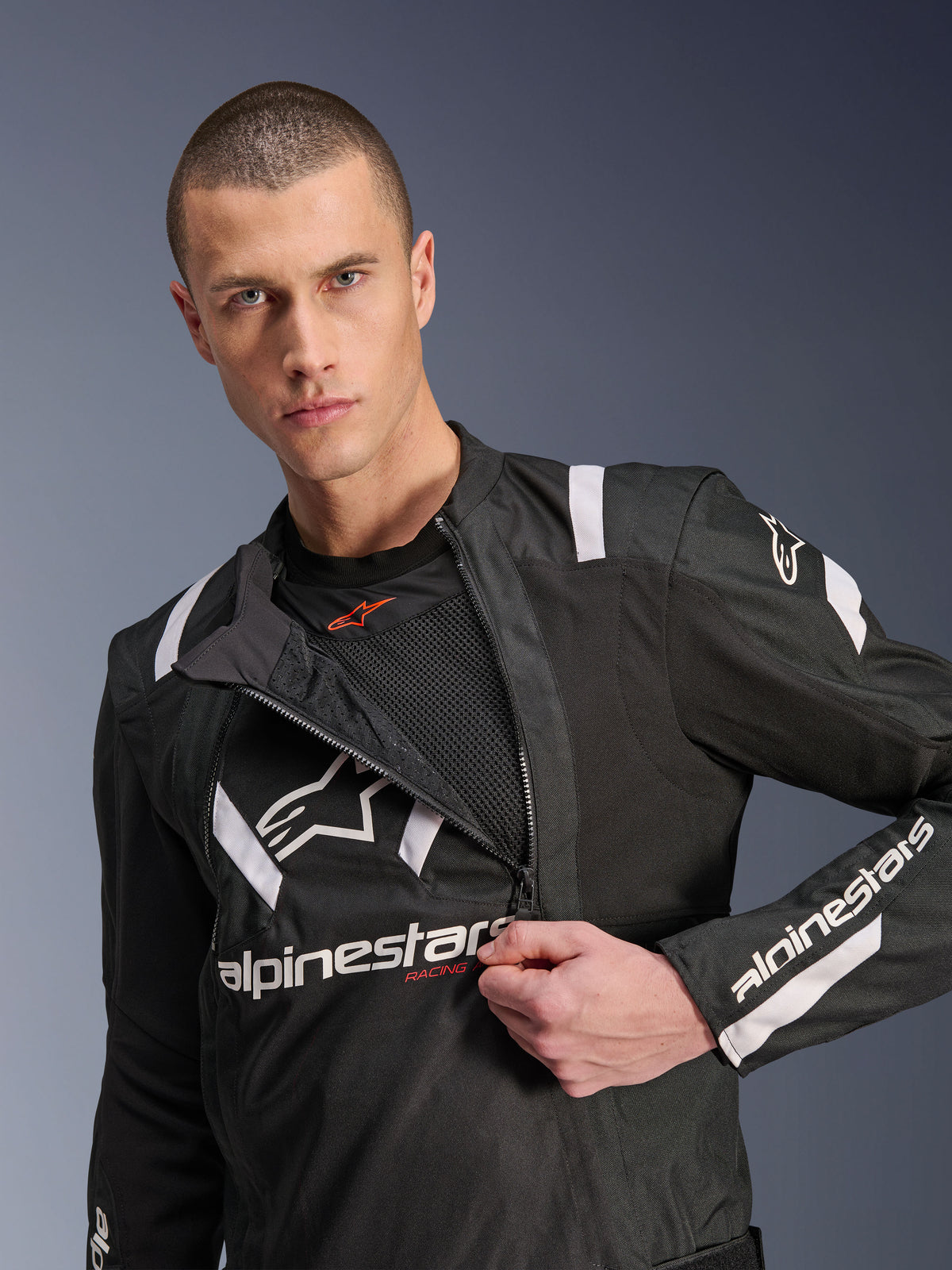 Alpinestars T-Stunt Air Jacket, svart och vit motorcykeljacka, manlig modell som visar en delvis öppnad modulär frontpanel som avslöjar mesh-lager för luftflöde, vita Alpinestars-logotyper på bröst och ärmar, sportig design för vägkörning