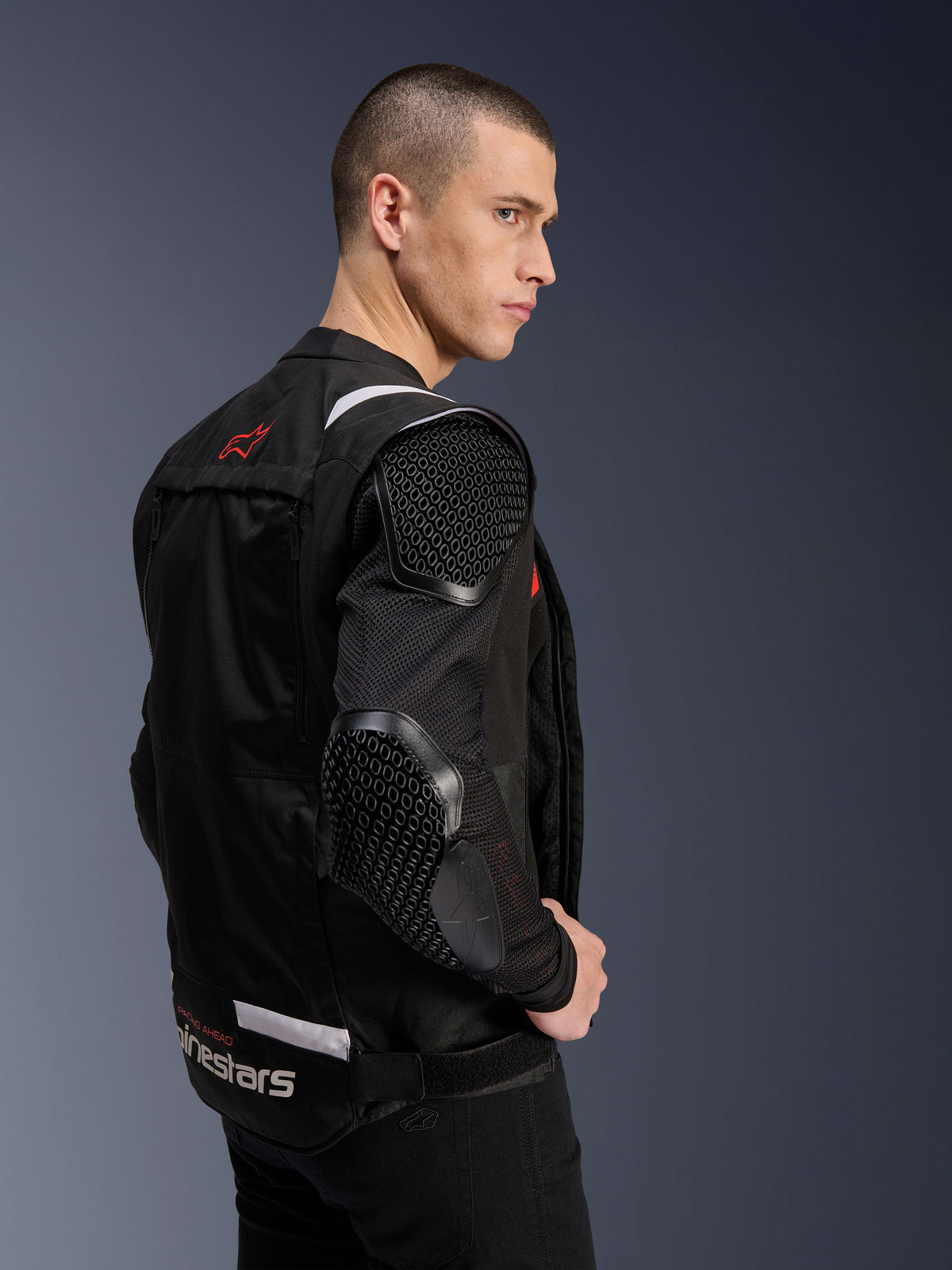 Alpinestars T-Stunt Air-jacka, motorcykeljacka, svart, sidoprofil av en modell som bär den sportigt skurna jackan med mesh-ärmar och synliga bikupemönstrade skydd på axel och armbåge, röd logotyp på ryggen, vitt varumärke i midjan.