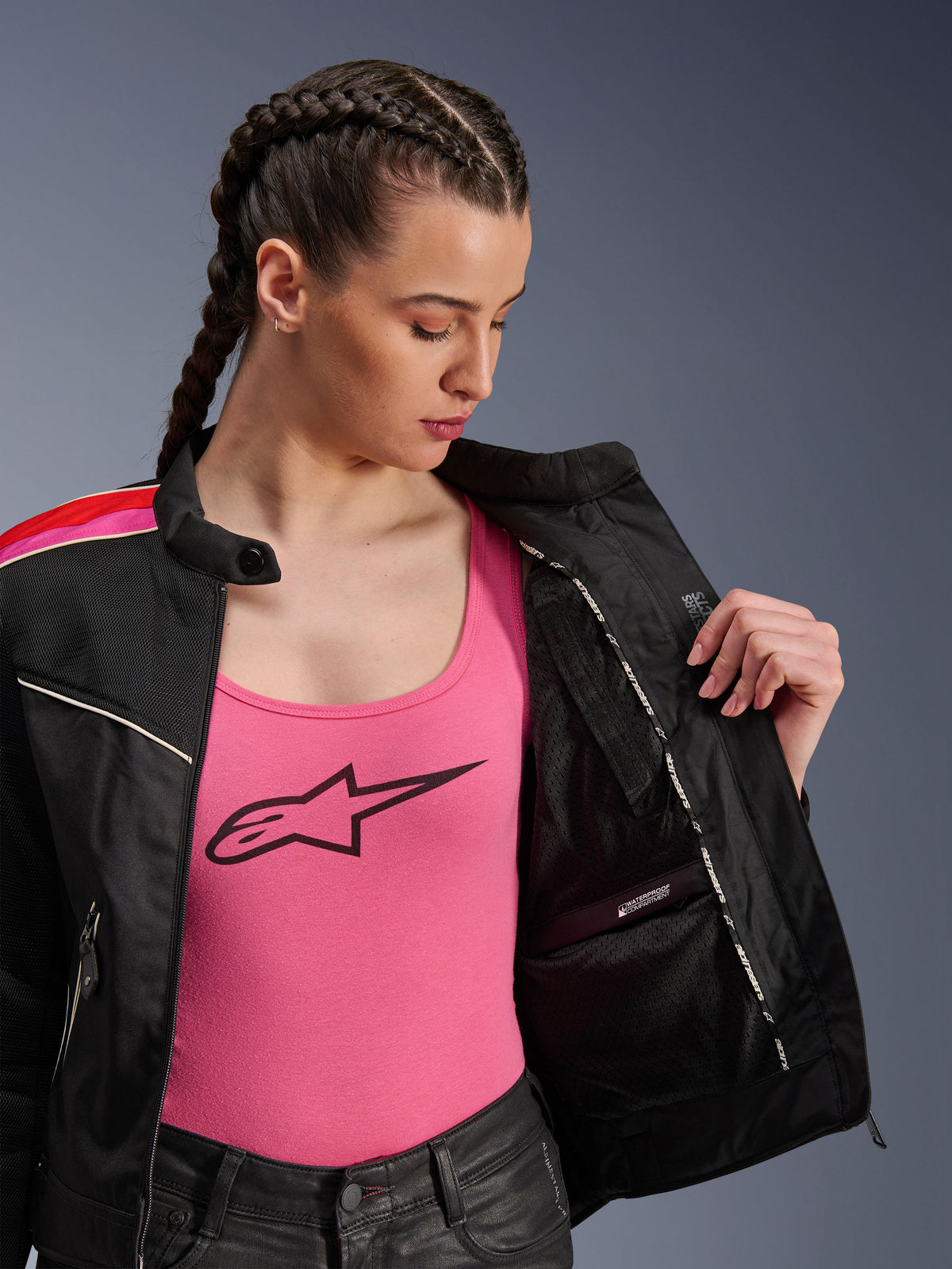 Kvinnlig förare i Alpinestars Stella T-Dyno Air-jacka i svart, klarröd och fuchsia, visas delvis öppen för att visa det inre vattentäta facket, bärs över ett rosa linne med logotyp, framhäver ventilerande meshpaneler och skräddarsydd biker-passform.