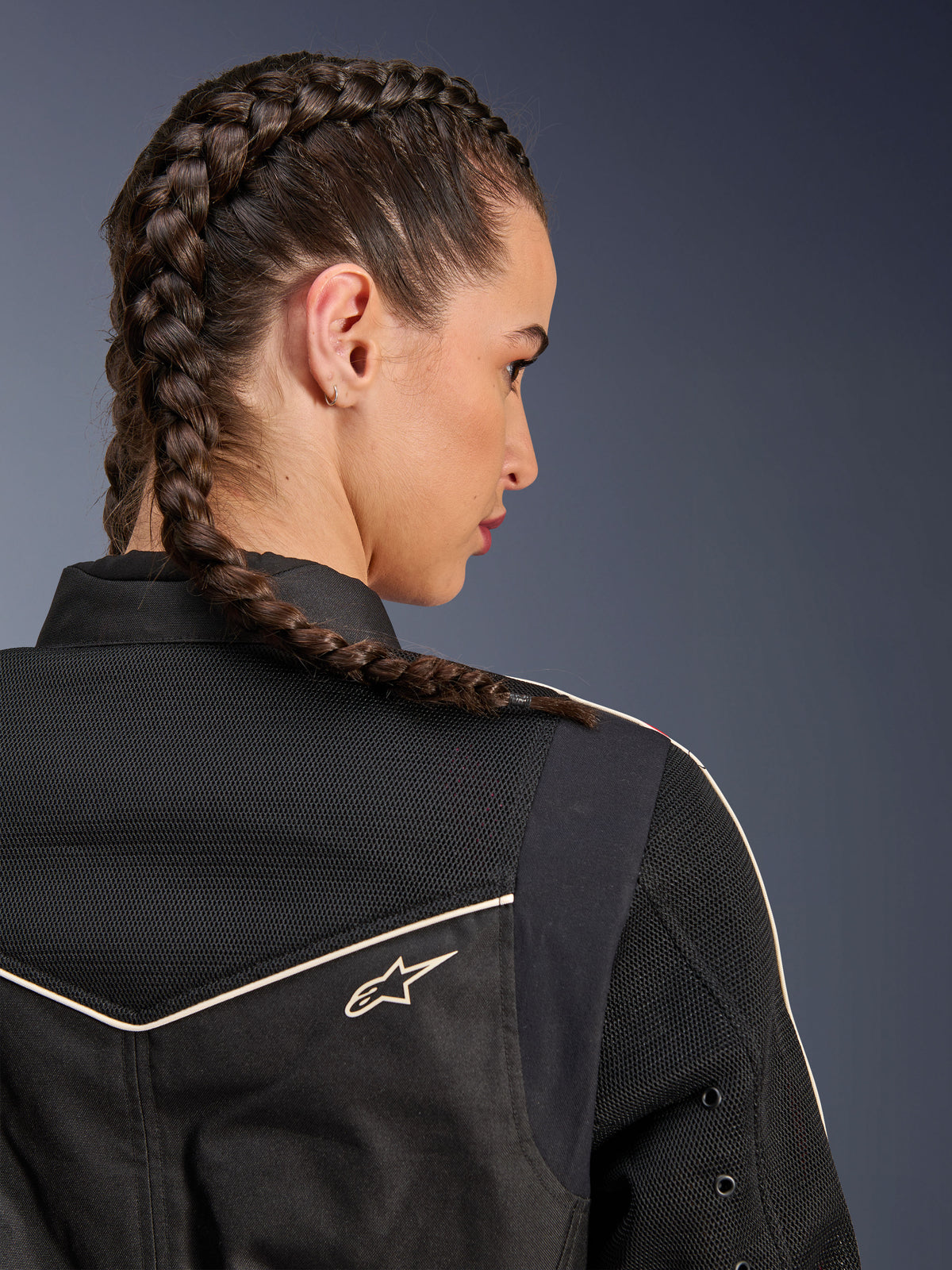 Kvinna som bär Alpinestars Stella T-Dyno Air Jacket, närbild bakifrån med fokus på axlar och ok, svart med ventilerande 3D-meshpaneler, vita passpoaler och en liten Astars-logotyp, modell med flätat hår, urbana motorcykelkläder