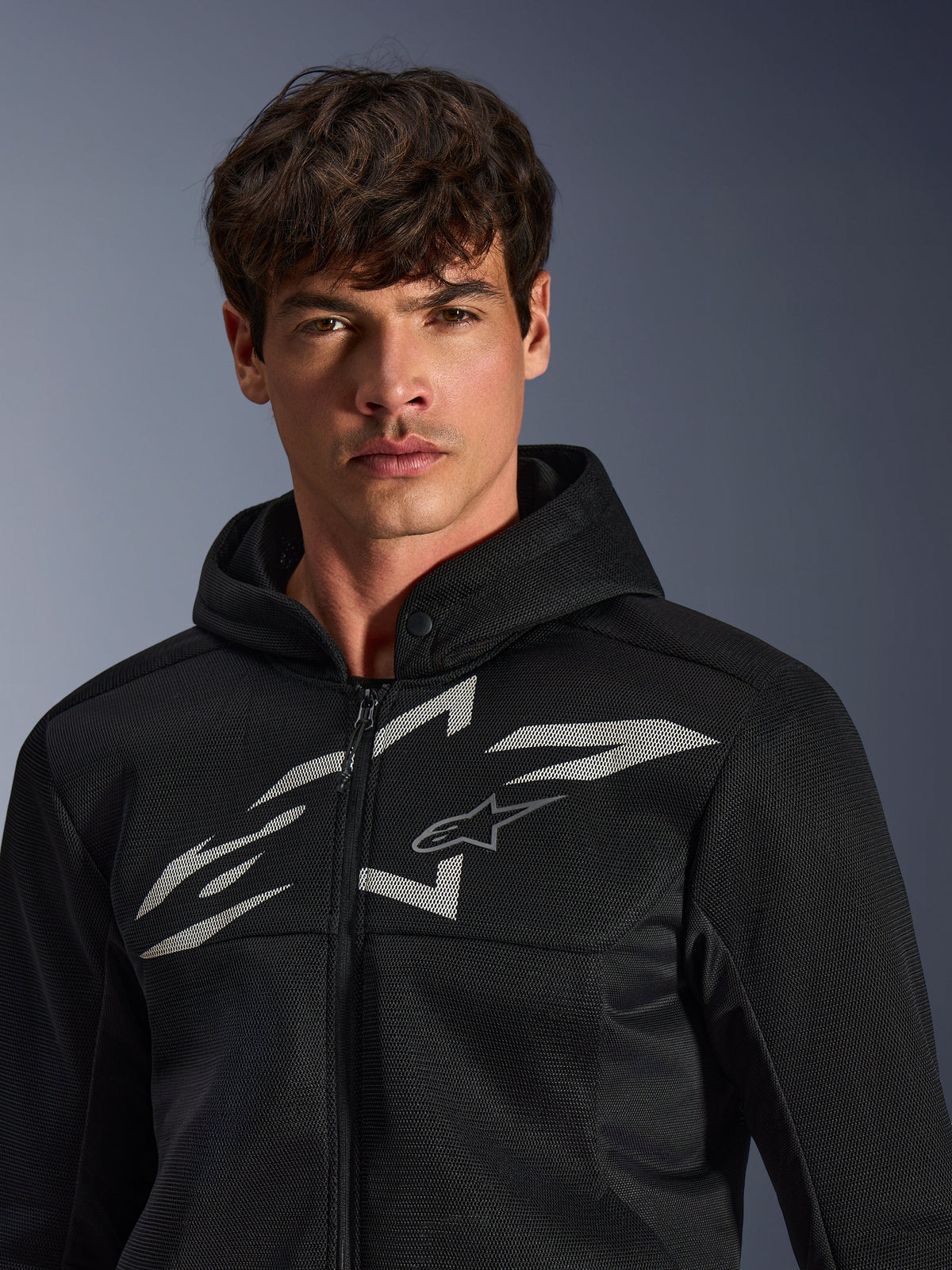 Alpinestars Chrome Superairflow Hoodie, svart motorcykelhoodie med 3D-mesh-konstruktion och en stor vit grafisk logotyp på bröstet, bärs av en modell, urban sportig körutrustning med modern ventilerad design.