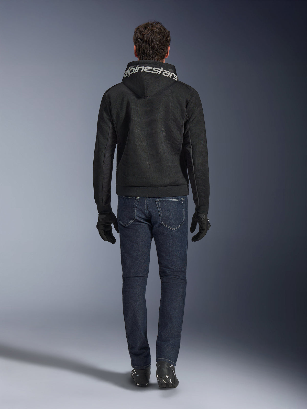 Alpinestars Chrome Superairflow Hoodie, svart motorcykelhoodie med vit logotyp på huvan, sedd bakifrån, bärs med mörkblå jeans, svarta handskar och svartvita stövlar, urban sportig körutrustning