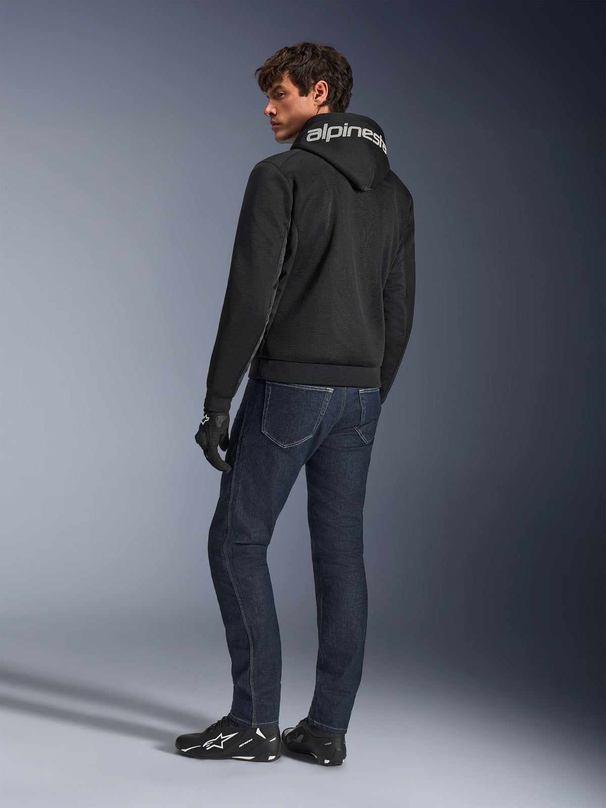 Alpinestars Chrome Superairflow Hoodie, svart, ryggvy på manlig modell, med vit Alpinestars-logotyp på huvan, bärs med mörka denimjeans och svarta handskar, urban design för motorcykelutrustning