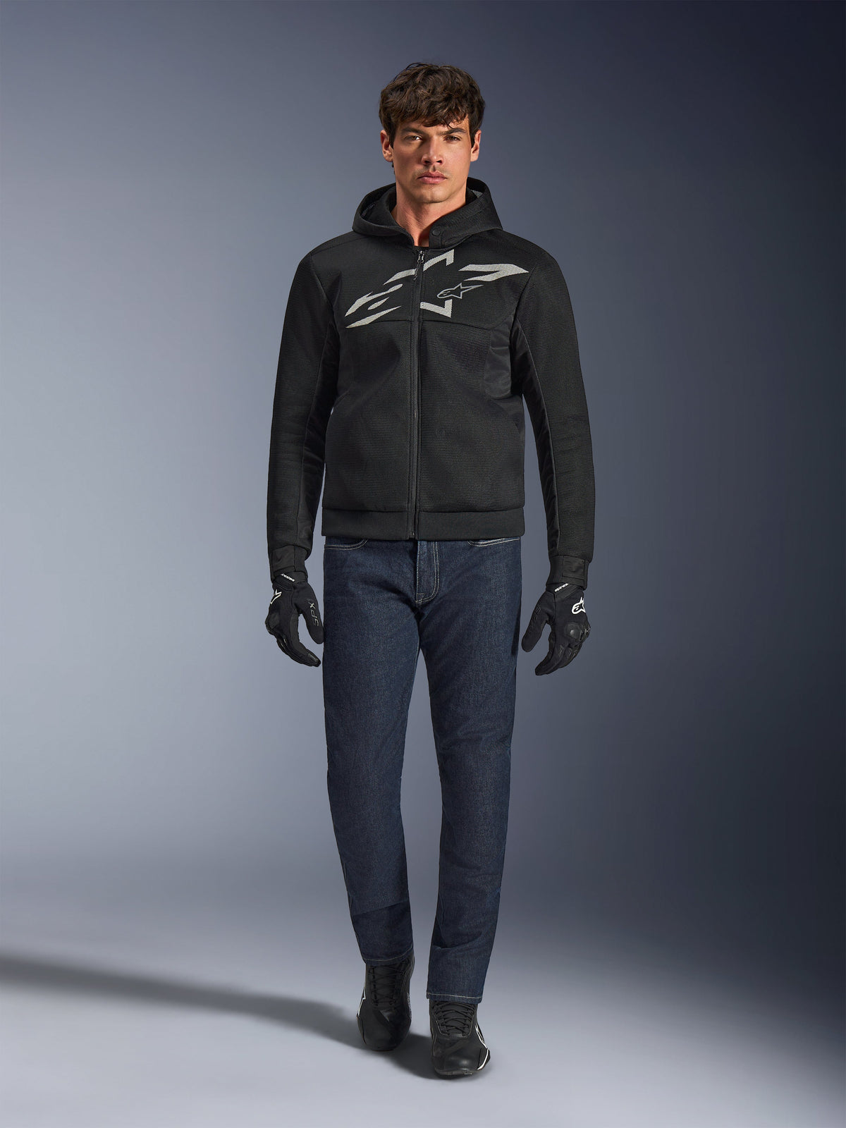 Motorcykelförare i Alpinestars Chrome Superairflow Hoodie i svart och grått, matchad med mörkblå jeans, svarta motorcykelhandskar och svarta skor. Hoodien har en stor stiliserad logotyp på bröstet och en 3D-meshstruktur för prestanda under höga temperaturer.