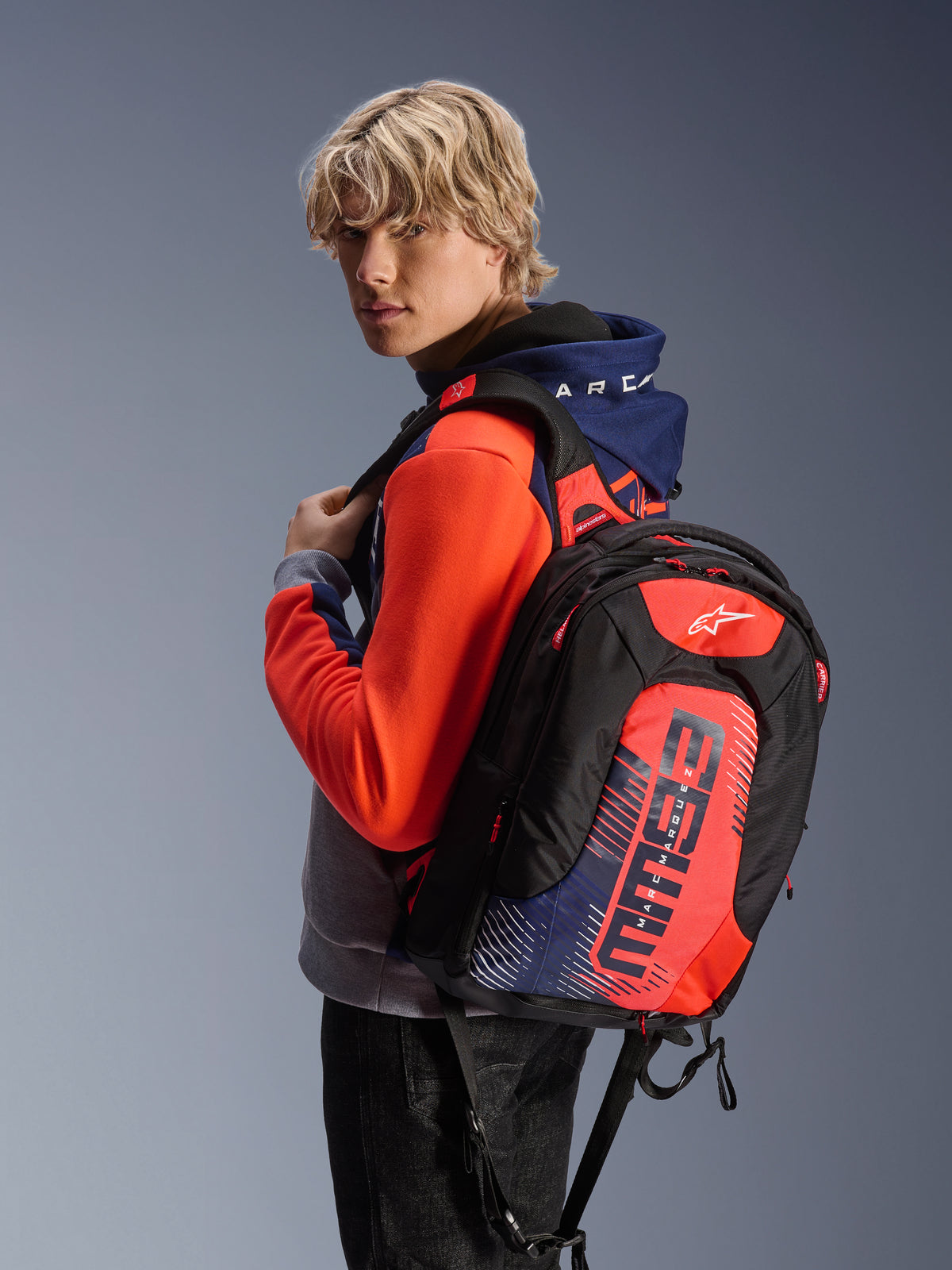 Alpinestars MM93 City Hunter V2 ryggsäck, svart med röd fluo och marinblå detaljer, med Marc Marquez MM93-signaturgrafik, bärs med en matchande sportig hoodie, smidig aerodynamisk profil för motorcykelpendling
