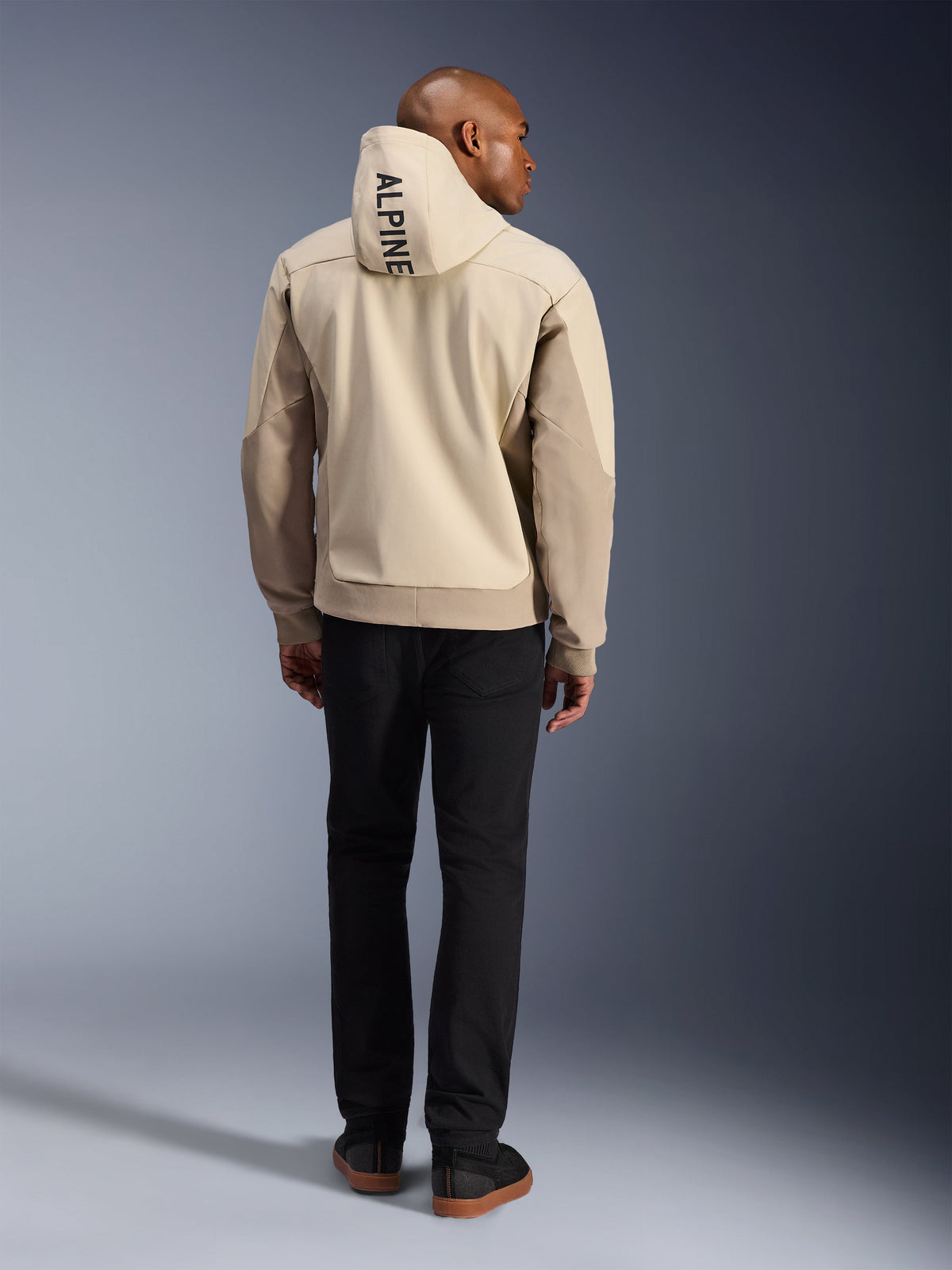 Alpinestars Radium Tech Hoodie, Mastic Aluminum, vy bakifrån som visar vertikal 'ALPINE'-text på huvan, teknisk 3L vindtät skaljacka för urban motorcykelkörning med beige paneler, buren med svarta byxor
