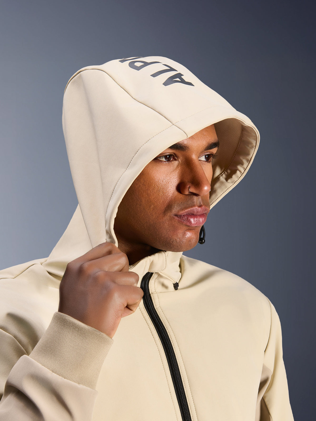 Alpinestars Radium Tech Hoodie i Mastic Aluminum, närbild på en man med huvan uppfälld, med Alpinestars-logotyp på toppen, urban motorcykeljacka med tekniskt vindtätt skal, modern streetwear-estetik för stadskörning