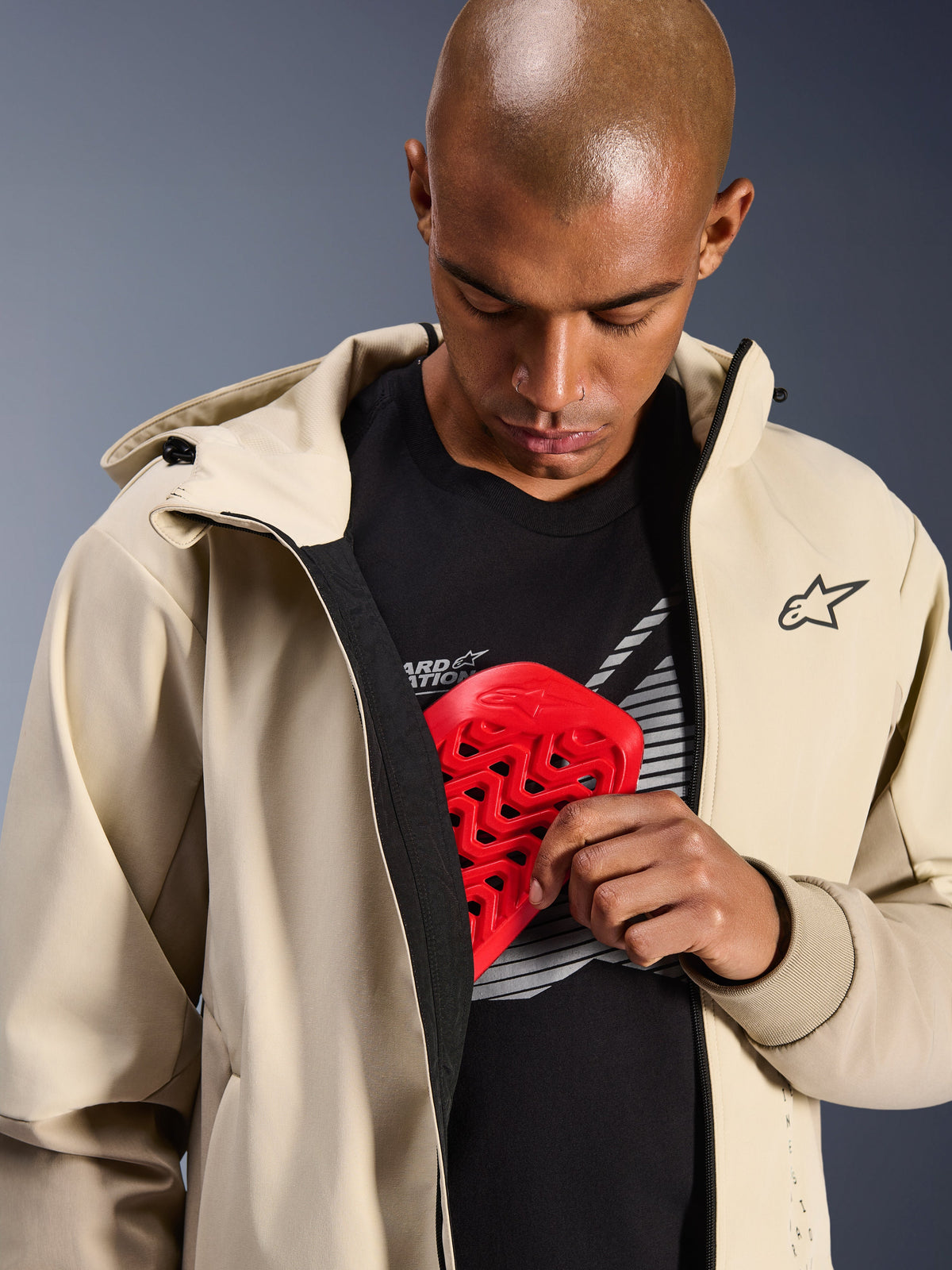 Alpinestars Radium Tech Hoodie i Mastic Aluminum, urban motorcykeljacka, modell som visar invändig skyddsficka med ett rött Nucleon bröstskydd, vindtätt tekniskt skal med en avslappnad hoodie-design och Alpinestars-branding.