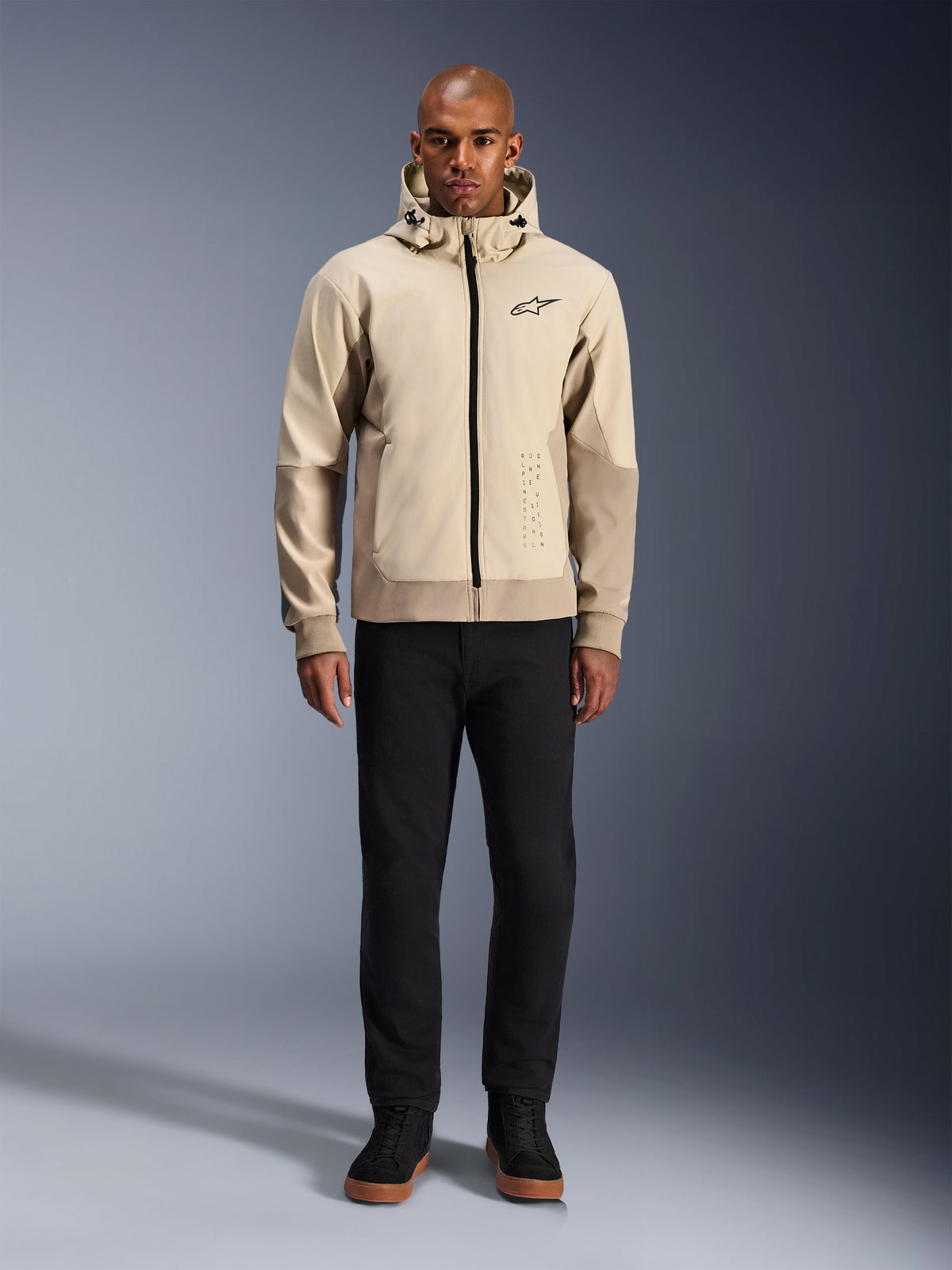 Alpinestars Radium Tech Hoodie i Mastic Aluminum, urban motorcykelhoodie som bärs av en manlig modell med svarta byxor och svarta kängor, med en genomgående svart dragkedja, liten logotyp på bröstet och atletisk passform för stadskörning.