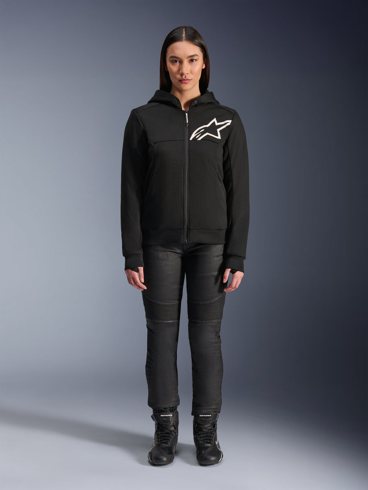 Kvinna som bär Alpinestars Stella Chrome V2 Sport Hoodie, svart med vit Astars-logotyp på bröstet, matchad med svarta tekniska körbyxor och stövlar, urban motorcykeldesign.
