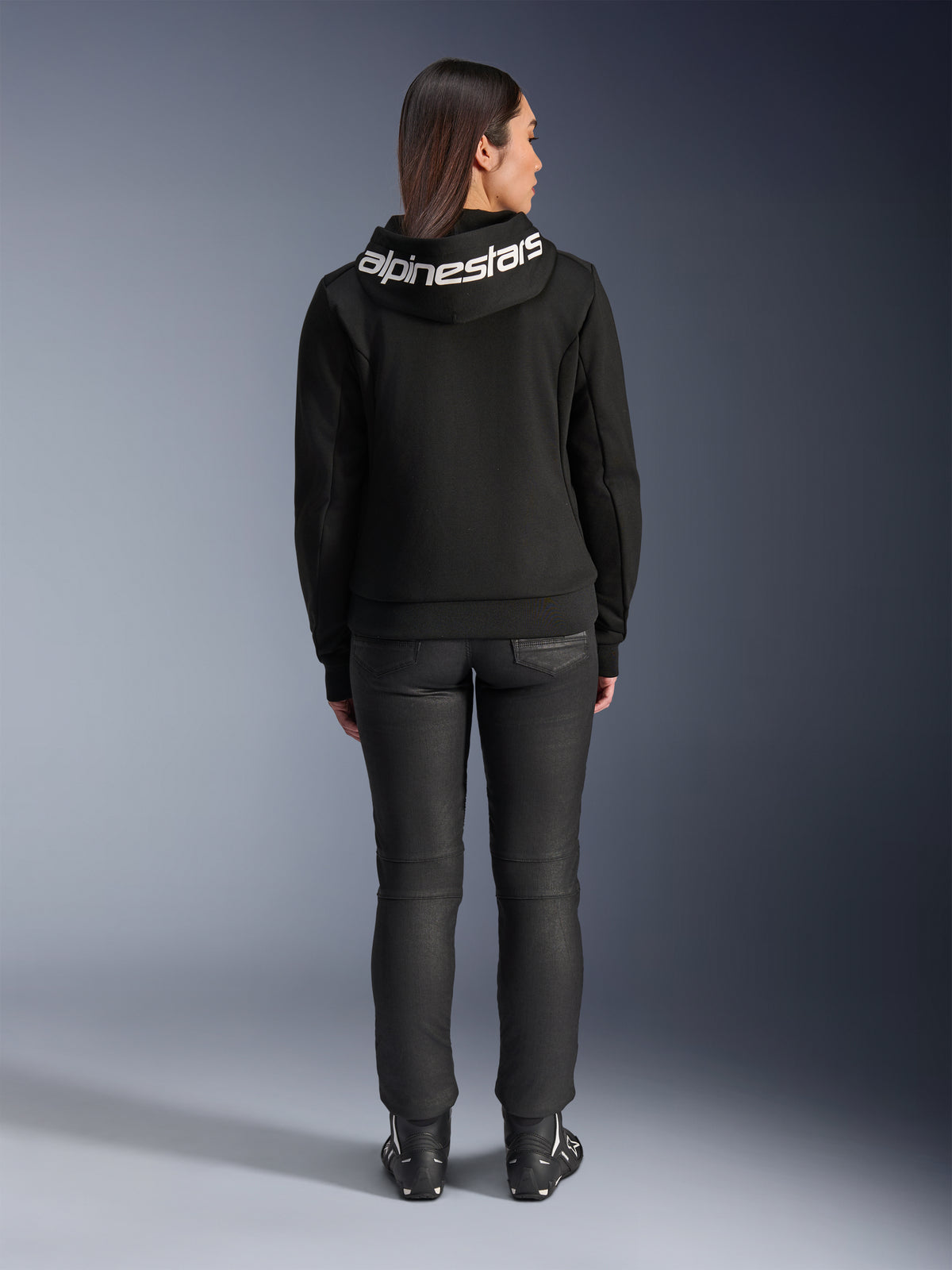 Vy bakifrån av en modell som bär Alpinestars Stella Chrome V2 Sport Hoodie i svart, med en framträdande vit Alpinestars-logotyp över huvan, matchad med svarta mc-byxor och motorcykelstövlar, urban sportmotorcykelutrustning för kvinnor