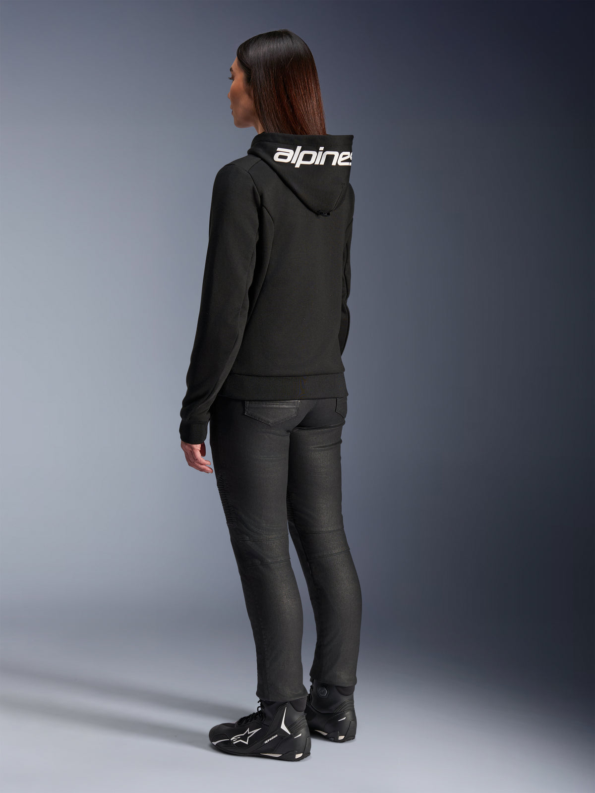 Kvinna i Alpinestars Stella Chrome V2 Sport Hoodie, sedd bakifrån, svart motorcykelhoodie med vit Alpinestars-logotyp på huvan, kombinerad med svarta tekniska mc-byxor och svarta motorcykelskor, avslappnad utrustning för urban pendling
