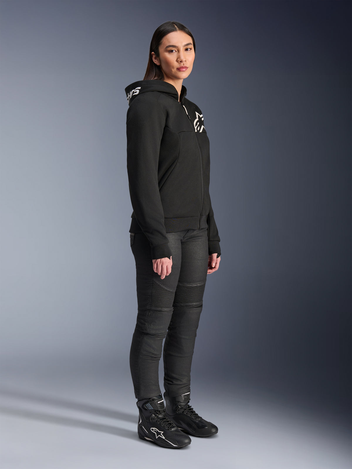 Kvinna som bär Alpinestars Stella Chrome V2 Sport Hoodie i svart, med vit Alpinestars-logotyp, kombinerad med svarta motorcykelbyxor och stövlar, urban mc-utrustning för kvinnor med en avslappnad atletisk passform