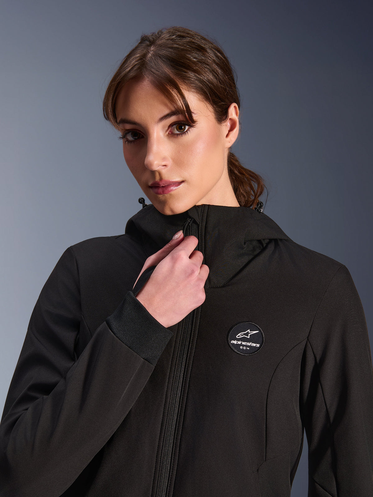 Woman Stella Radium Tech Hoodie, Black, worn by a woman adjusting the front zipper, featuring circular Alpinestars logo patch on the chest, ribbed stretch cuffs, and windproof textile construction for urban riding