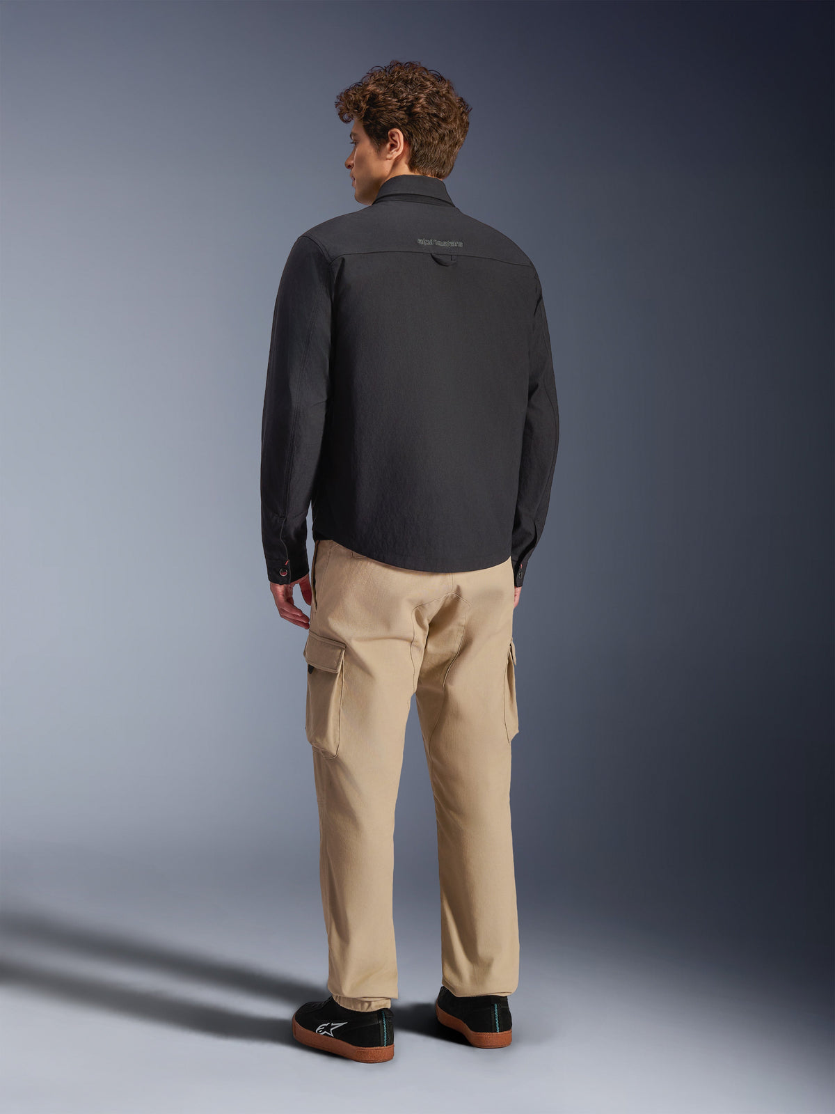 Alpinestars Bryon WR överskjorta, svart, sedd bakifrån på modell, kombinerad med beige cargobyxor och svarta sneakers, urban motorcykeljacka med vaxad canvas-textur och ren minimalistisk ryggdesign