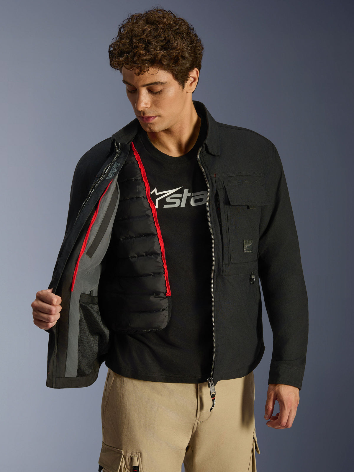 Alpinestars Bryon WR överskjorta, svart, urban motorcykeljacka som visas öppen för att visa en avtagbar svart kviltad termoväst med röda detaljer, bärs med beige cargobyxor och en t-shirt med logotyp, funktionell design inspirerad av arbetskläder