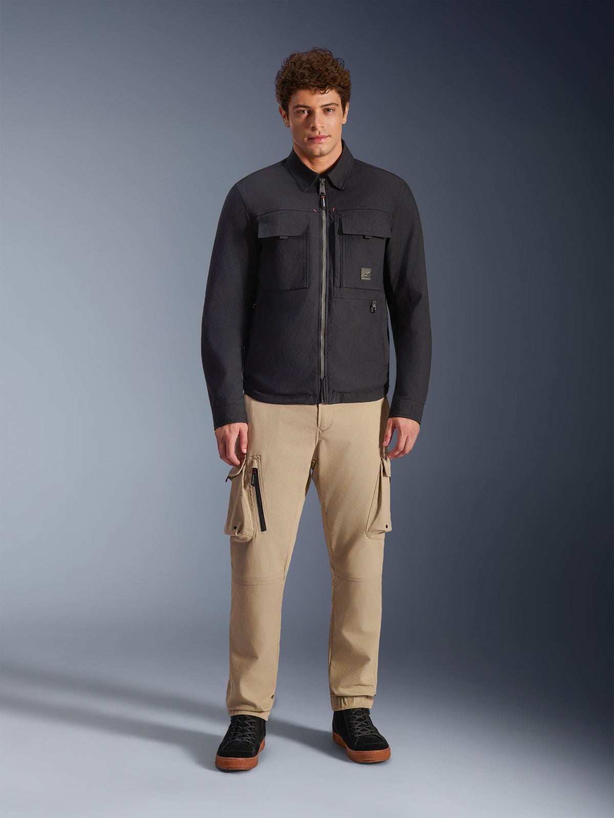 Manlig modell som bär Alpinestars Bryon Water Resistant Overshirt i svart, med bröstfickor och central dragkedja, stylad med beige cargobyxor och svarta sneakers, helkroppsporträtt i studiomiljö.