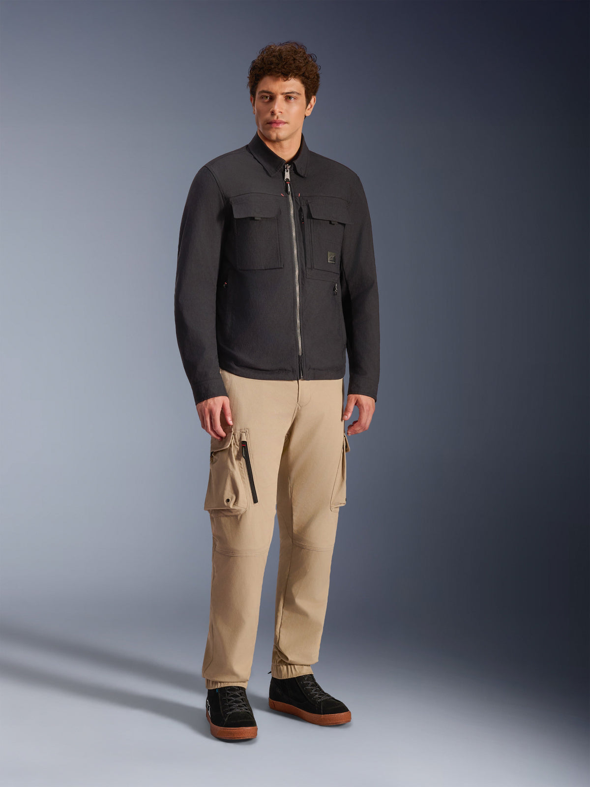 Alpinestars Bryon Water Resistant Overshirt, svart, bärs av manlig modell med khakifärgade cargobyxor och svarta skor, urban motorcykeljacka med arbetsklädesinspirerad design, helkroppsbild i studiomiljö