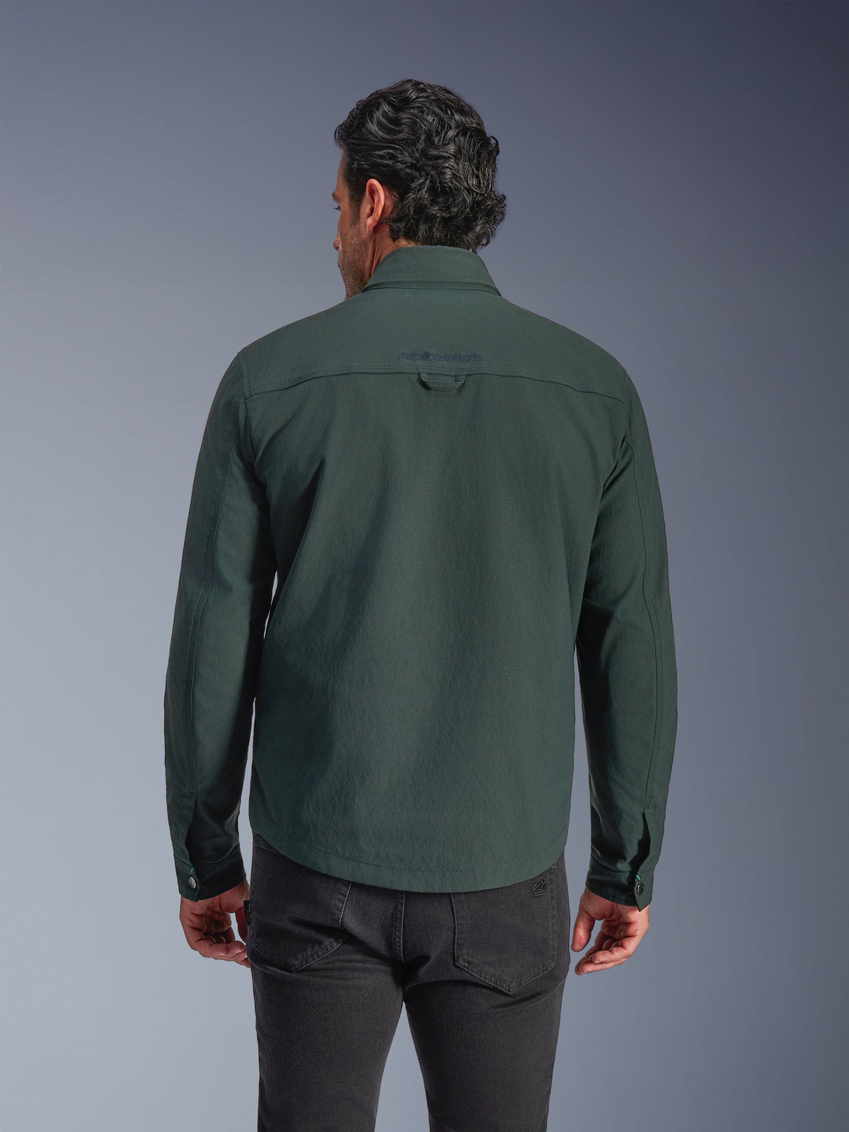 Alpinestars Bryon vattenavvisande överskjorta, mörk smaragdgrön, sedd bakifrån på modell, broderad logotyp på övre ryggen, slitstark vaxad canvas, urban motorcykeljacka kombinerad med mörka byxor