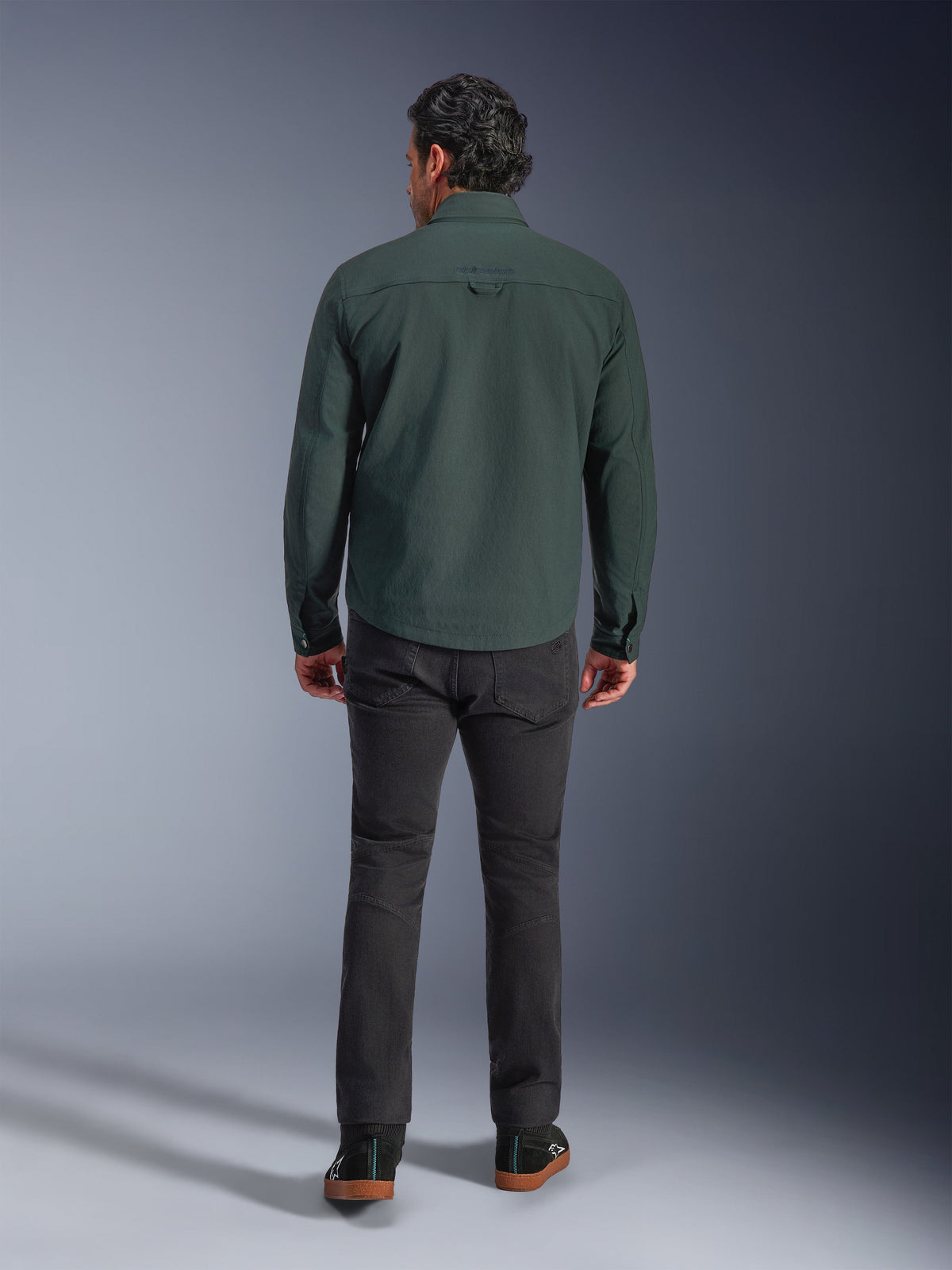 Alpinestars Bryon Water Resistant Overshirt i Dark Emerald, sedd bakifrån på modell, kombinerad med mörkgrå motorcykelbyxor och svarta körskor, visar en klassisk arbetsklädesinspirerad design med en något längre rygg för täckning vid körning.