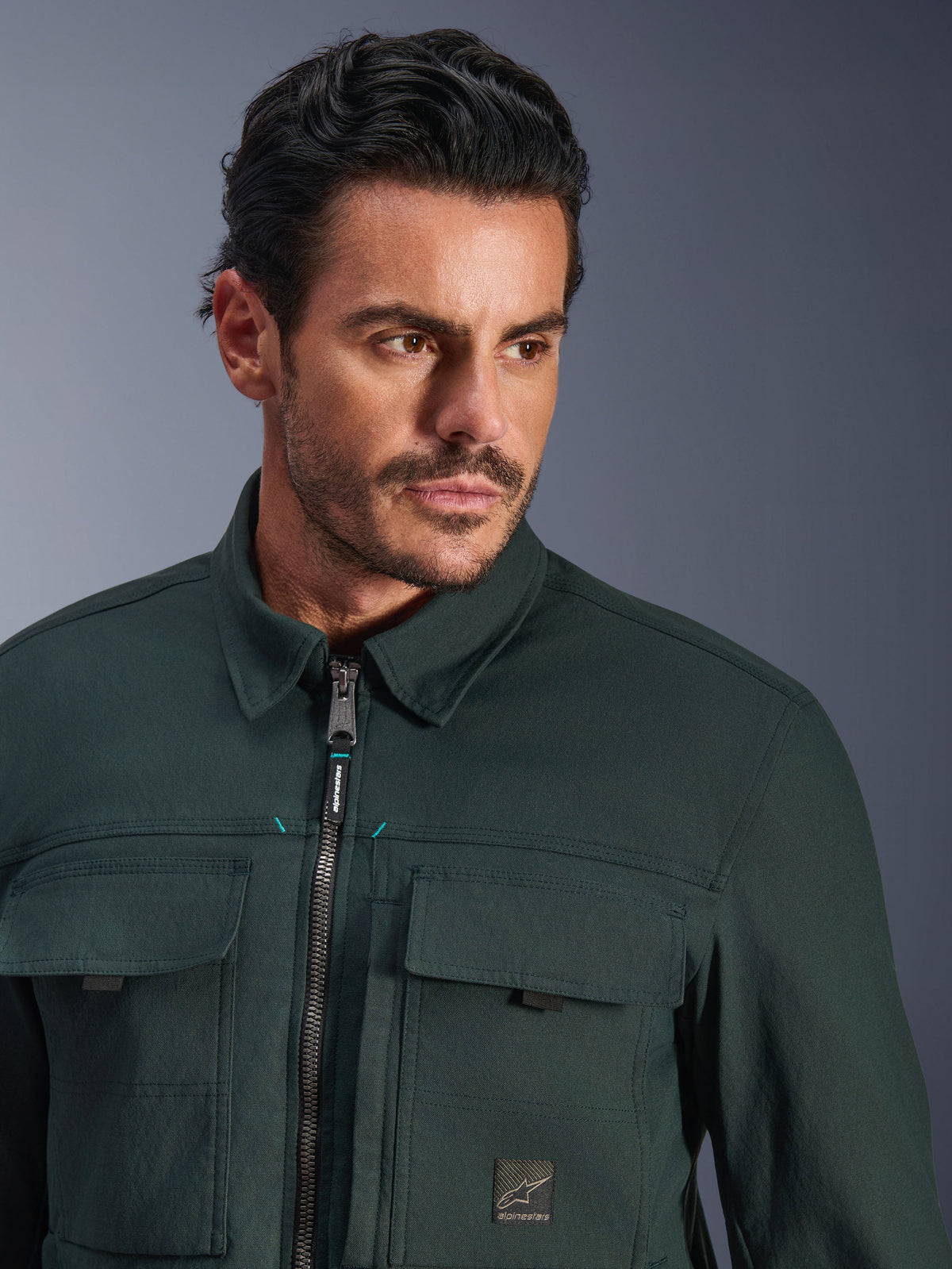 Alpinestars Bryon Water Resistant Overshirt i Dark Emerald, närbildsporträtt av en man som bär den urbana motorcykeljackan, med vaxad canvastextur, främre dragkedja med subtila teal-detaljer, cargofickor på bröstet och märkeslogotyp.