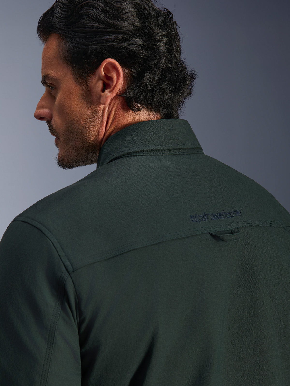 Alpinestars Bryon Water Resistant Overshirt i mörkt smaragdgrönt, närbild på övre delen av ryggen och kragen på en manlig modell, broderad Alpinestars-logotyp och en liten hängögla i tyg, tillverkad av texturerad vattenavvisande vaxad canvas för urban motorcykelkörning.