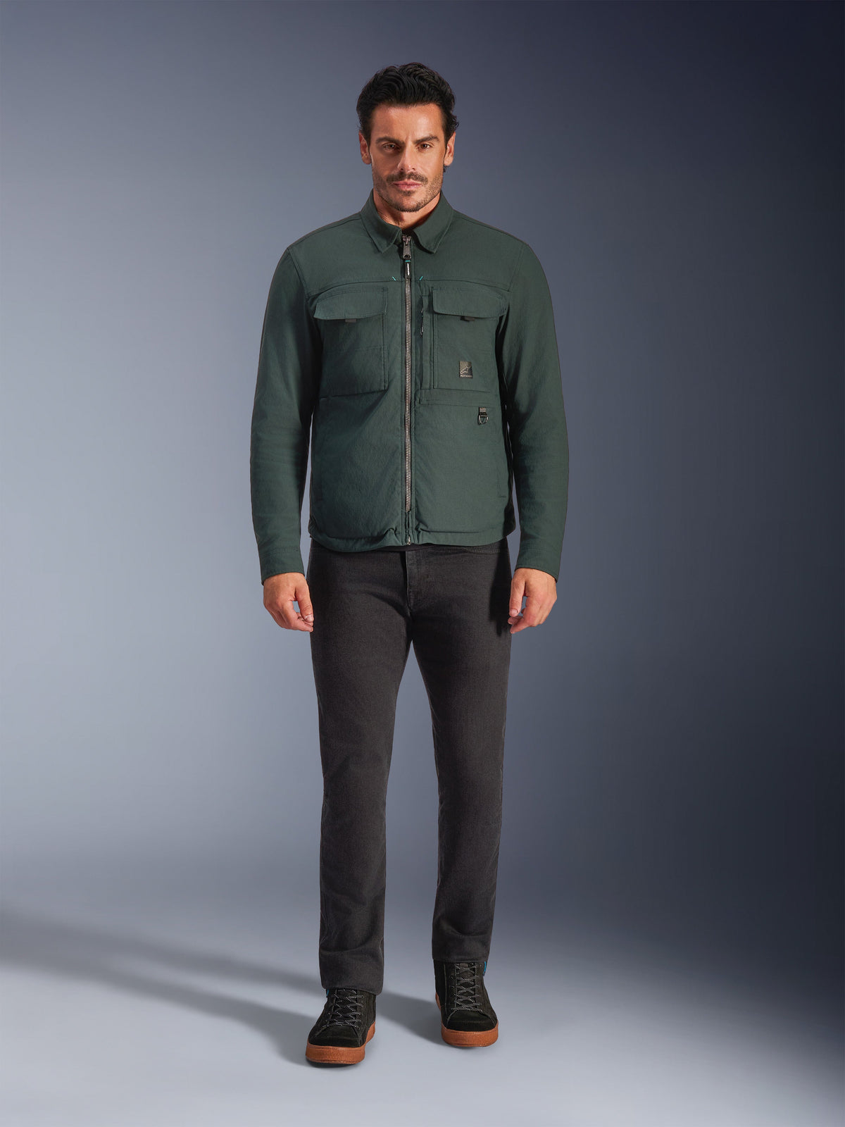 Alpinestars Bryon WR Overshirt, vattenavvisande motorcykeljacka, Dark Emerald, bärs med svarta byxor och svarta kängor, med vaxad canvas-konstruktion, cargofickor på bröstet och en modern urban design.