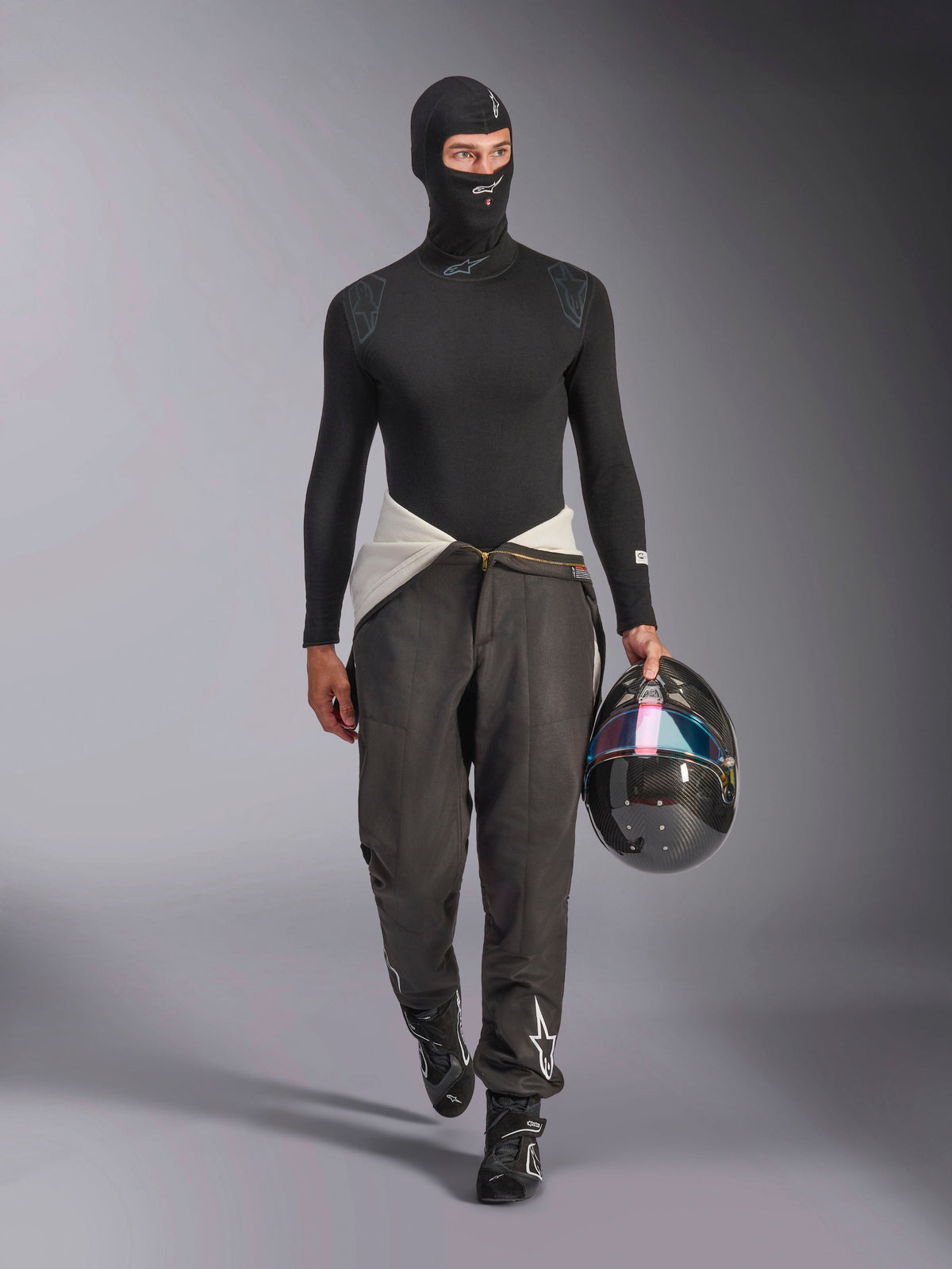 Alpinestars ZX Evo V3 Balaclava, Black, worn by a model in a full racing base layer outfit including a matching long-sleeve top and grey racing pants, holding a carbon fiber helmet with an iridescent visor, professional auto racing gear