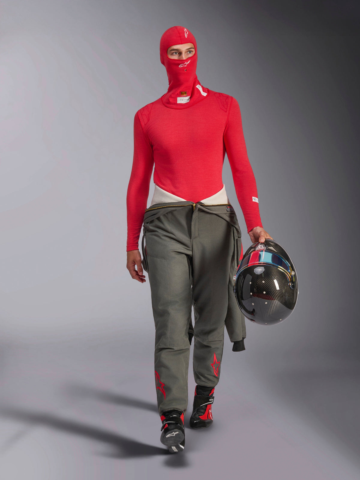 Alpinestars ZX Evo V3 Balaclava in Red, worn by a racing driver with a matching red long-sleeved base layer and grey racing pants, holding a carbon fiber helmet, professional flame-resistant auto racing gear