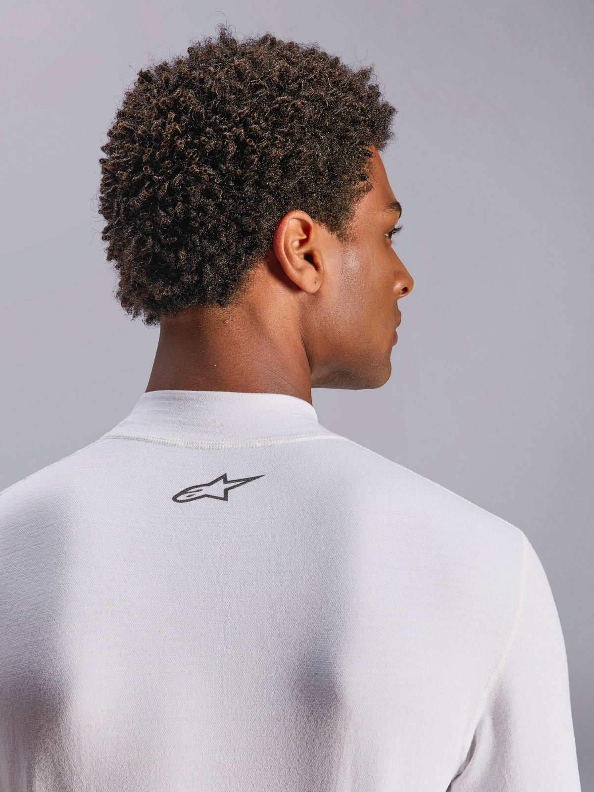 Alpinestars ZX Evo V3 Top, långärmat underställ för bilsport, vit, närbild av nackpartiet med svart Alpinestars-logotyp, tekniskt flamskyddat material, bärs av modell med mörkt lockigt hår