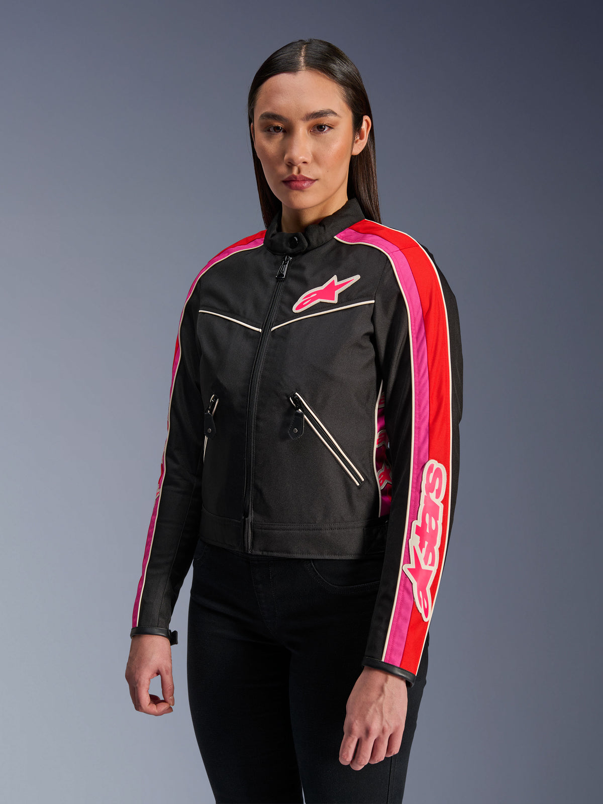 Kvinna som bär Alpinestars Stella T-Dyno WR-jacka i Black Bright Red Fuchsia, textil motorcykeljacka med vintageinspirerad biker-passform, livfulla fuchsiafärgade och röda ränder på ärmarna och en logotyp på bröstet, matchad med svarta byxor.