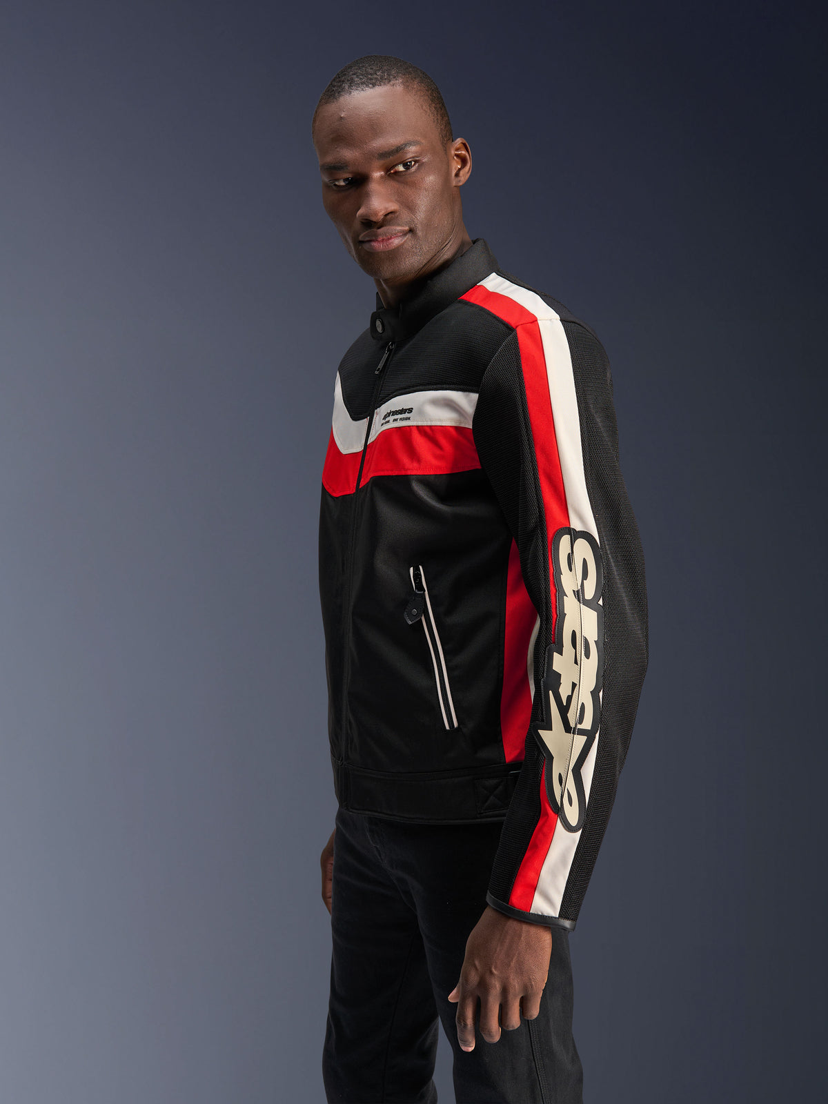 Manlig modell iförd Alpinestars T-Dyno Air-jacka i svart, klarrött och ecru, en vintageinspirerad mc-jacka i textil med 3D-meshpaneler, markerade röda och off-white ränder samt en tydlig Alpinestars-logotyp på ärmen.